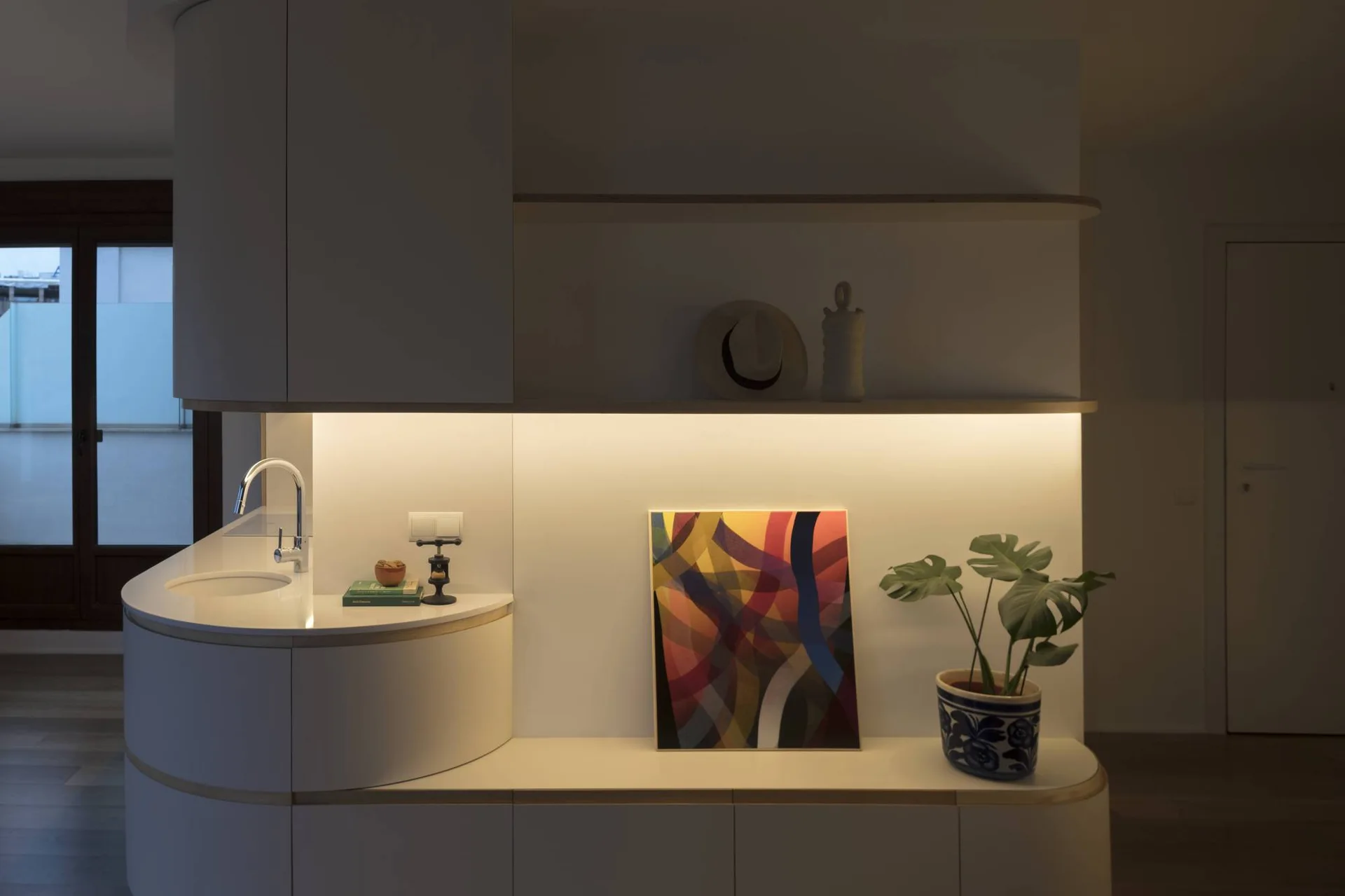 Minimalist white cabinetry with curved edges features an integrated sink, floating shelves with warm under-shelf lighting, and decorative artwork.