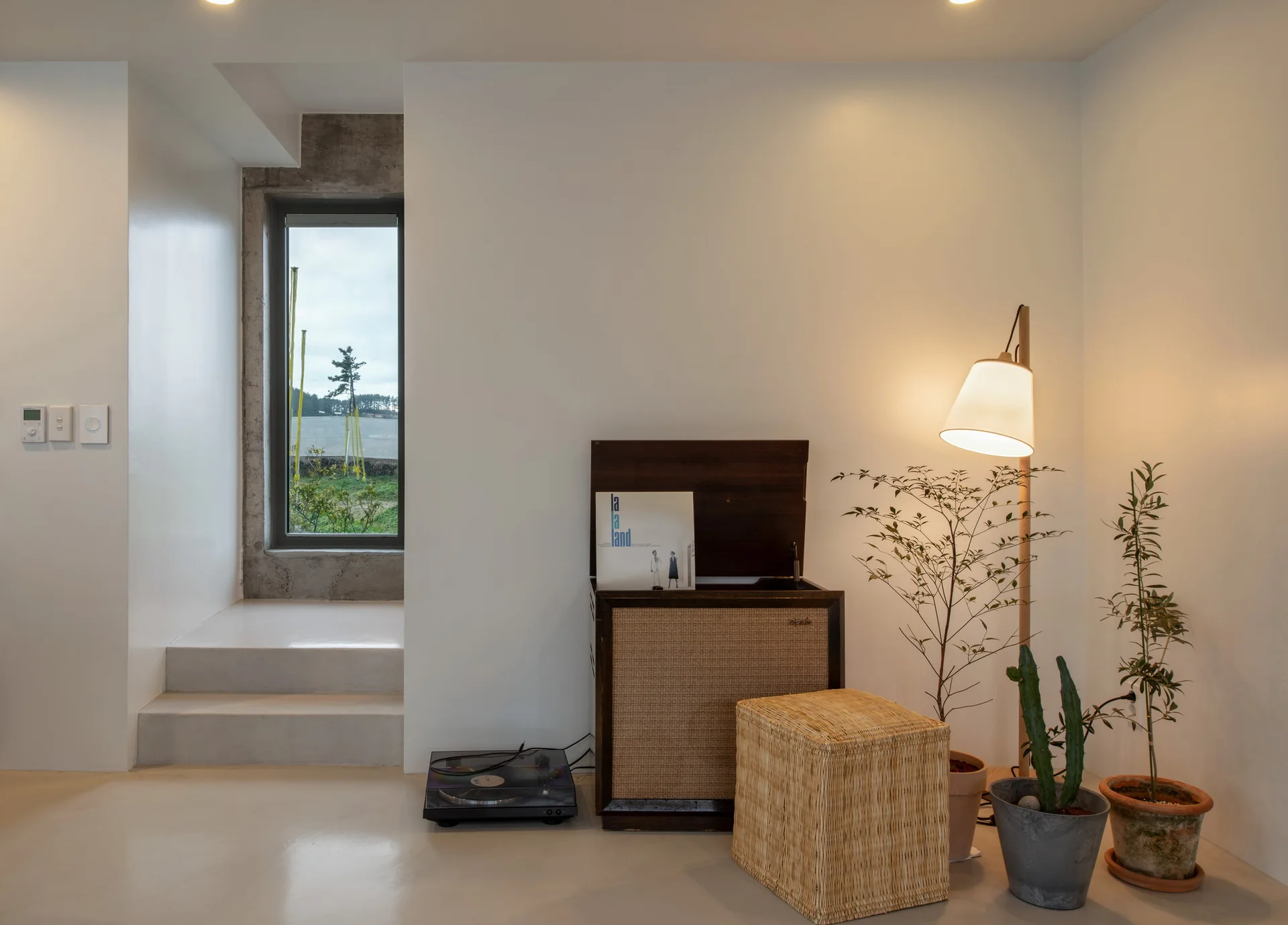 A vintage wooden record player stands against a white wall next to a warm floor lamp, a woven rattan stool, and potted plants.