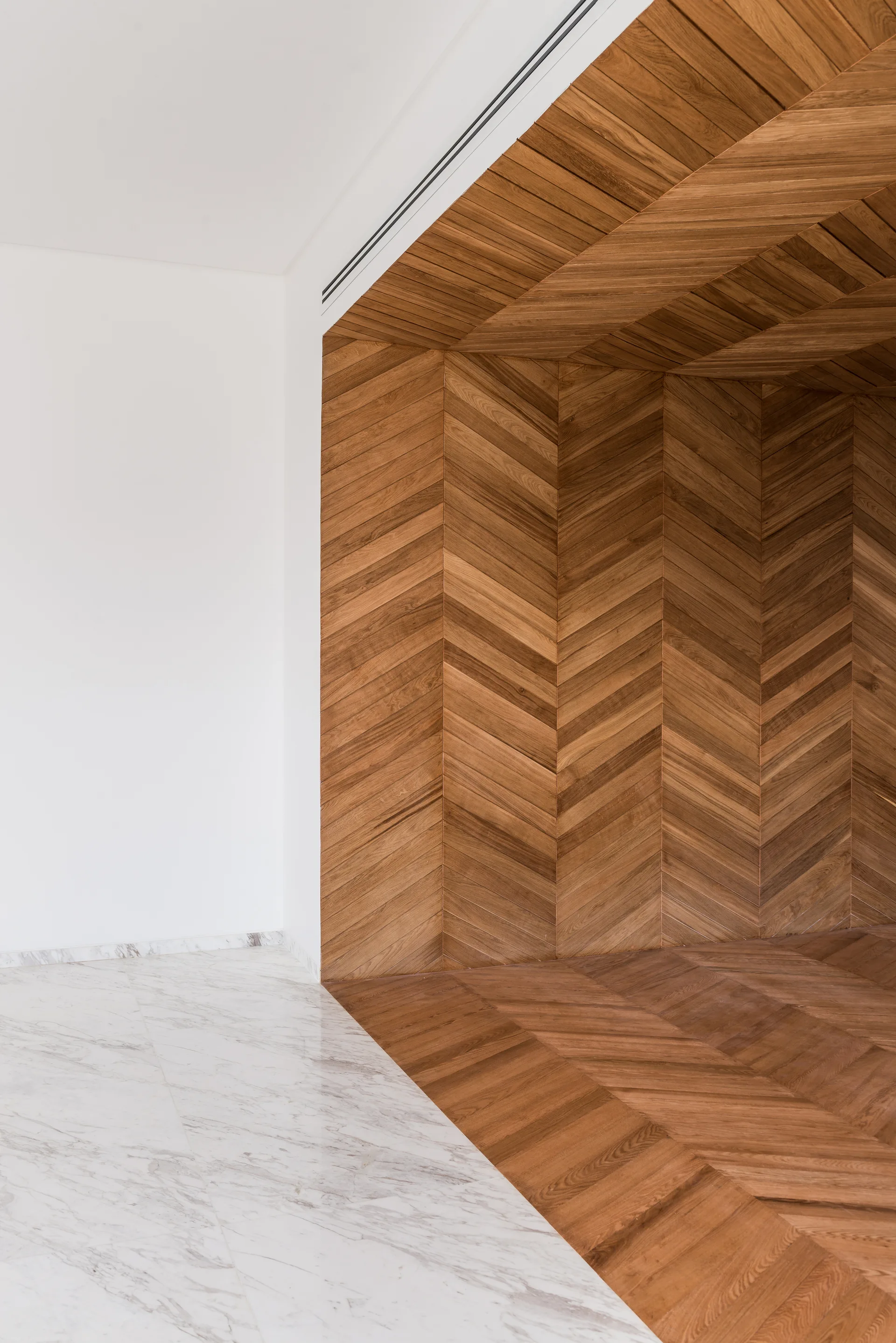A modern interior corner featuring white walls and marble tile flooring transitioning to a recessed area wrapped in warm wood herringbone paneling.