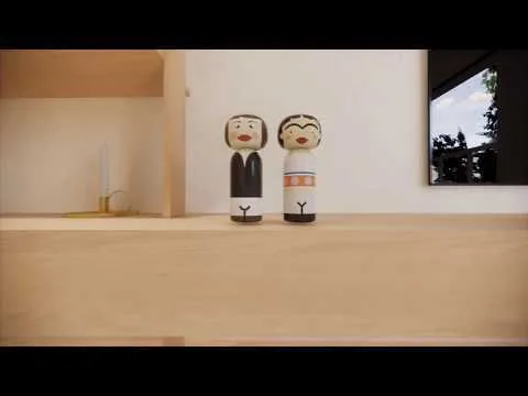 Two painted wooden kokeshi-style dolls sit on a light wood shelf next to a brass candlestick against a white wall.