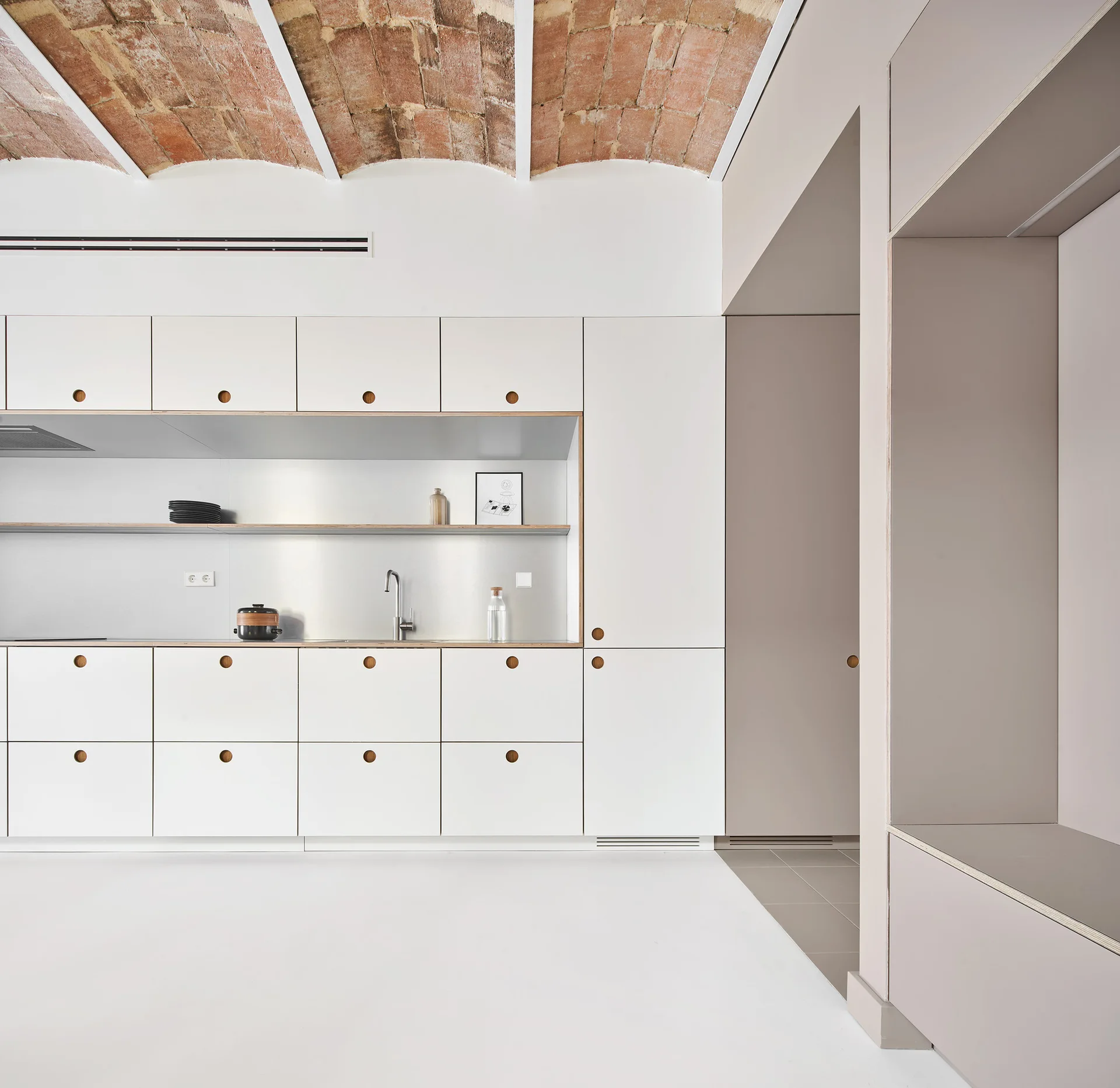 A minimalist white kitchen features flat-panel cabinets, a recessed stainless steel backsplash, and an exposed red brick vaulted ceiling.