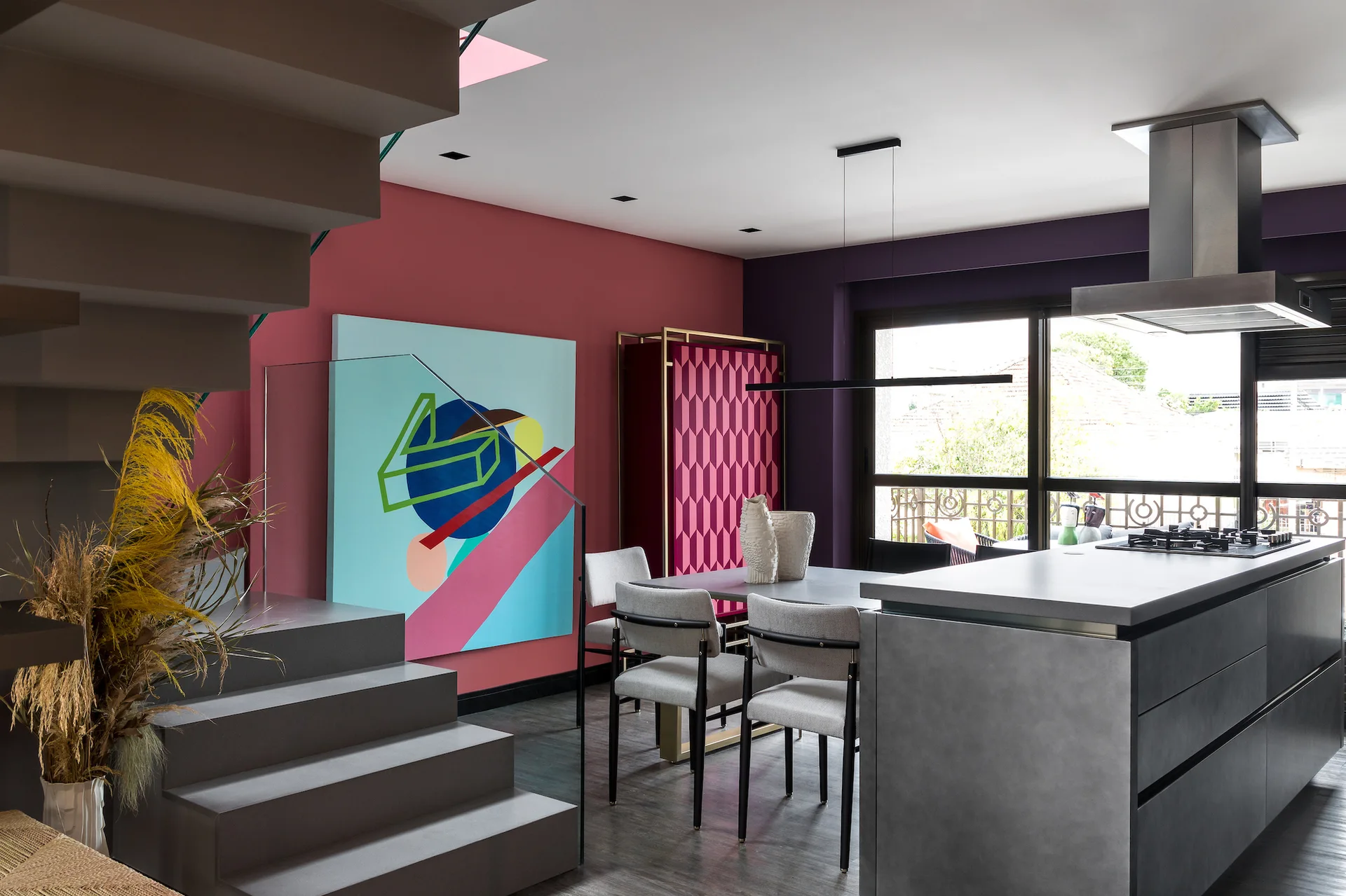 A modern dining room and kitchen features a grey island, bold pink and purple walls, a geometric patterned cabinet, and an abstract painting.