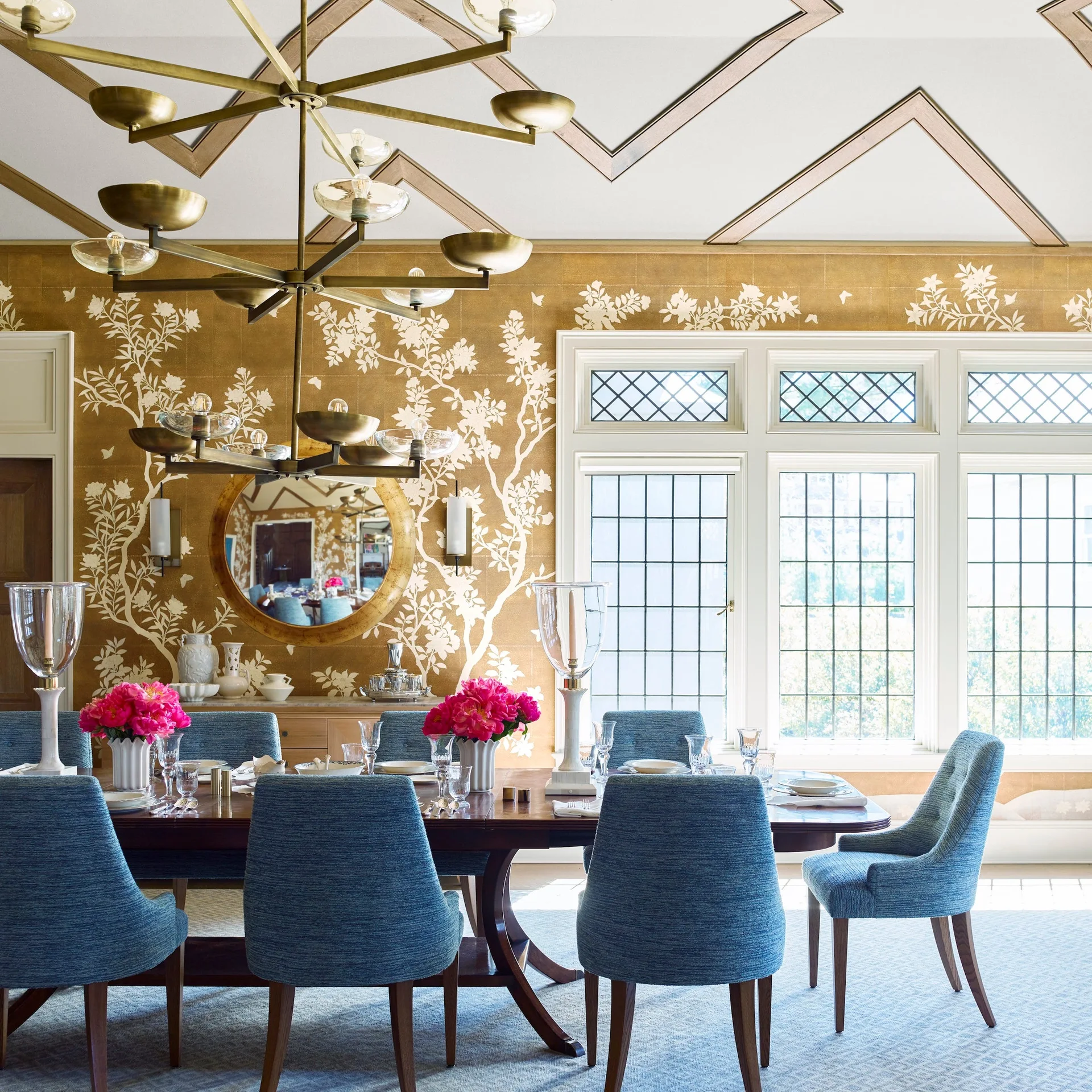 A formal dining room features blue upholstered chairs around a dark wood table, set against gold floral wallpaper and a geometric ceiling design.