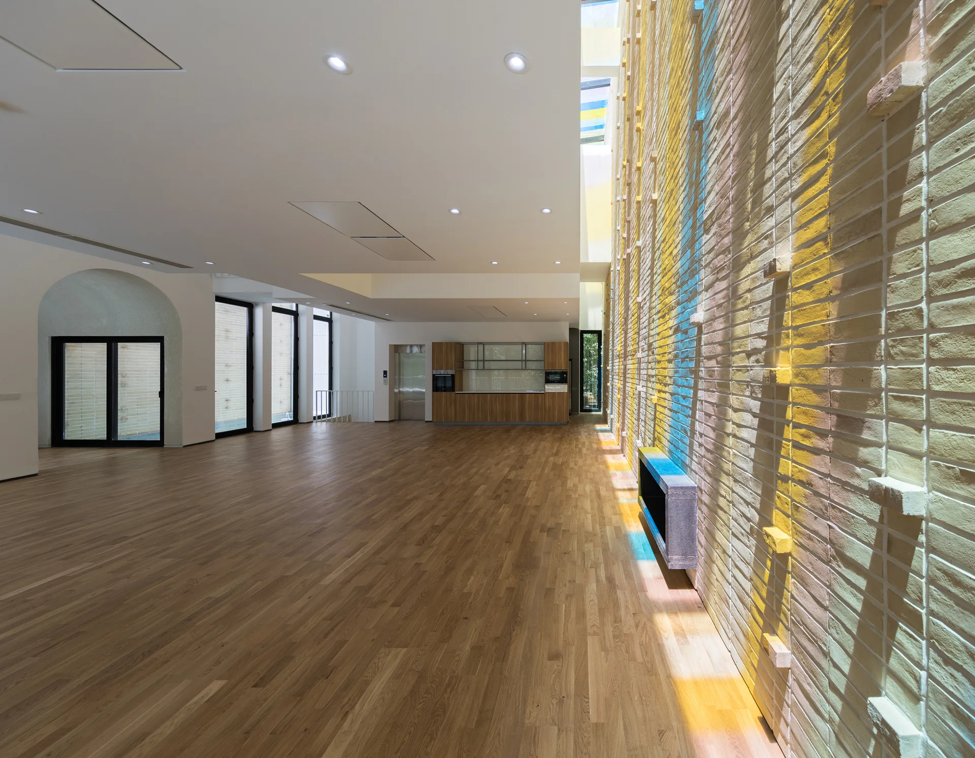 A minimalist open-plan interior featuring natural wood floors, an arched entryway, and a long brick wall illuminated by colored skylight reflections.