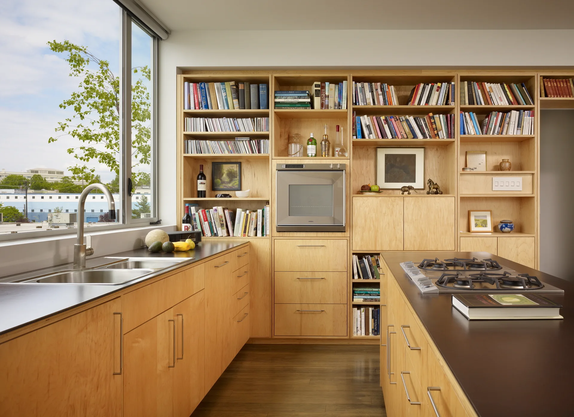 A modern kitchen featuring light wood cabinetry, integrated open bookshelves filled with books, dark countertops, and a central island with a gas cooktop.