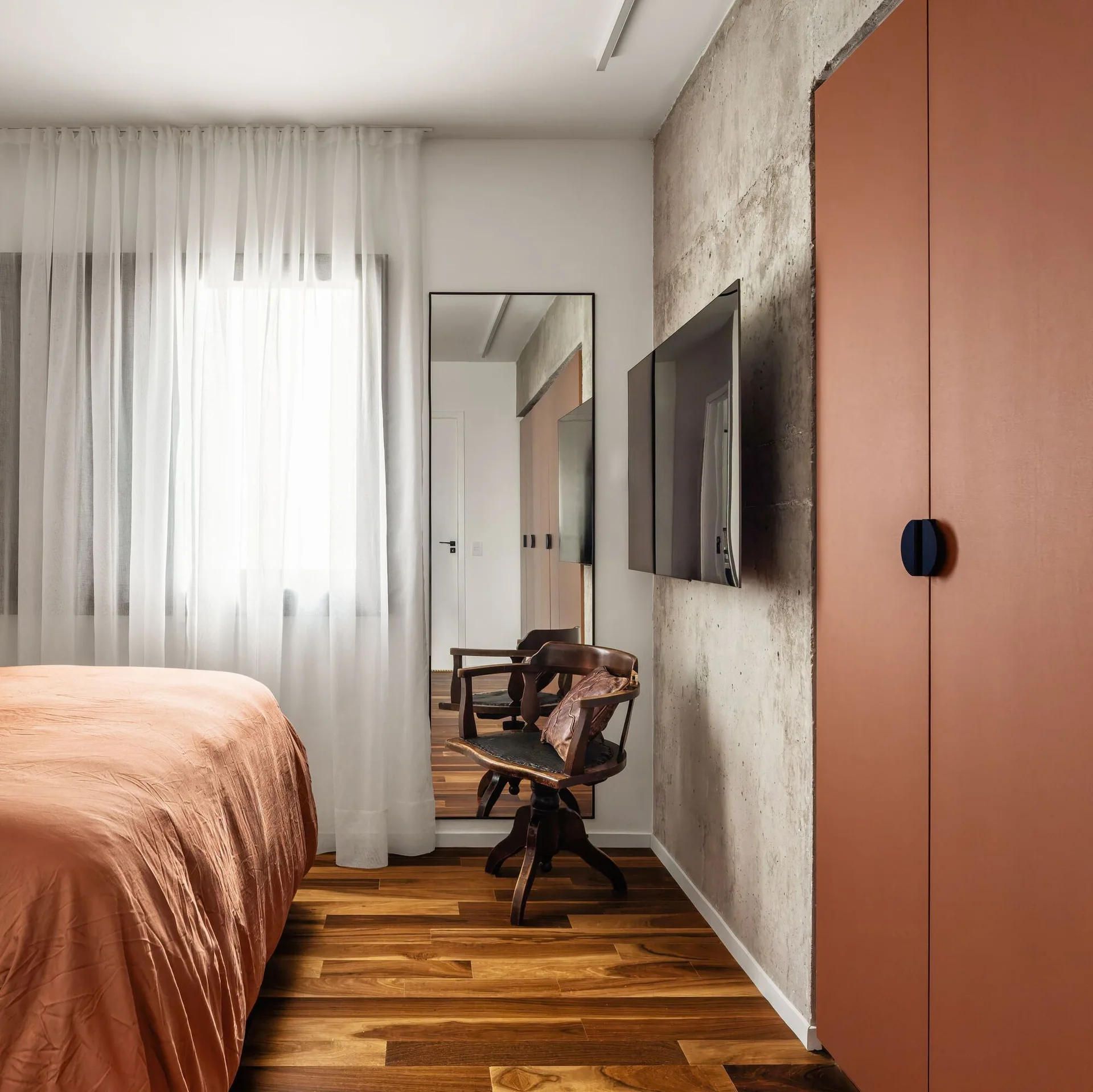 A modern bedroom featuring an exposed concrete wall, a terracotta wardrobe, a vintage wood chair, and a bed with rust-colored linens.