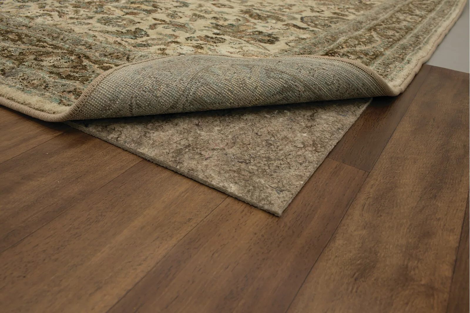 The corner of an ornate patterned area rug is folded back to show a thick grey felt rug pad underneath on dark wood flooring.