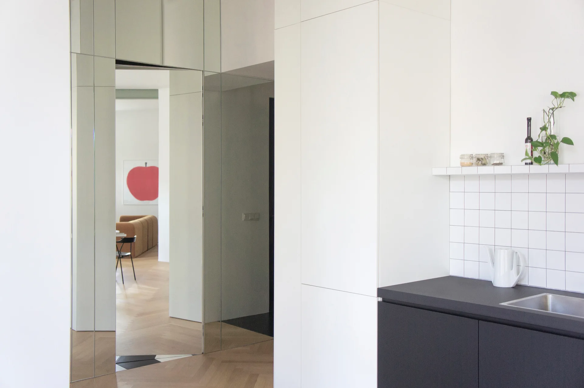 A minimalist kitchen features matte black base cabinets, a white tiled backsplash, and a mirrored wall surrounding a doorway to an adjacent room.