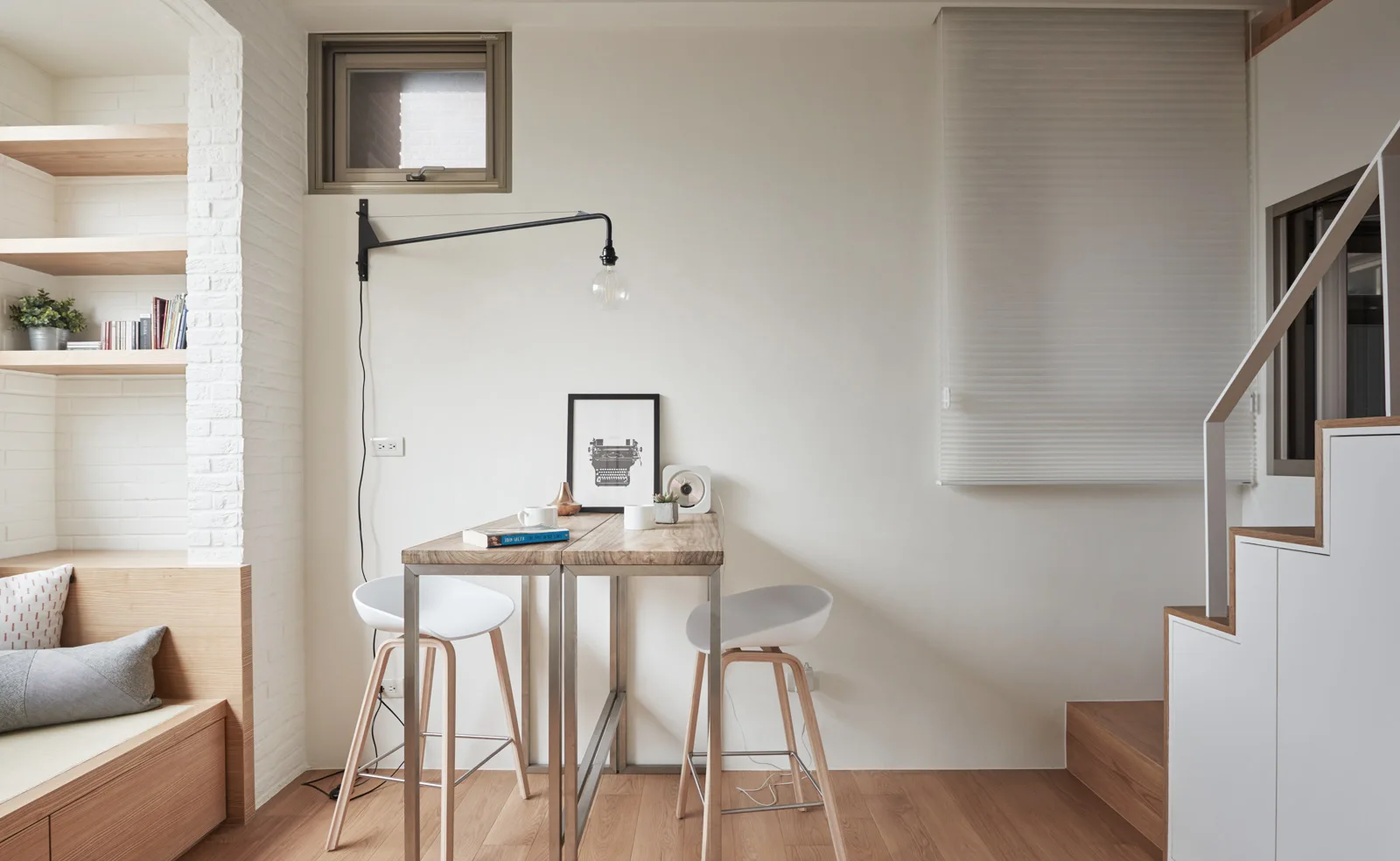 A compact dining nook features a high wooden table with two white stools, a black wall-mounted lamp, and a white staircase with wood steps.