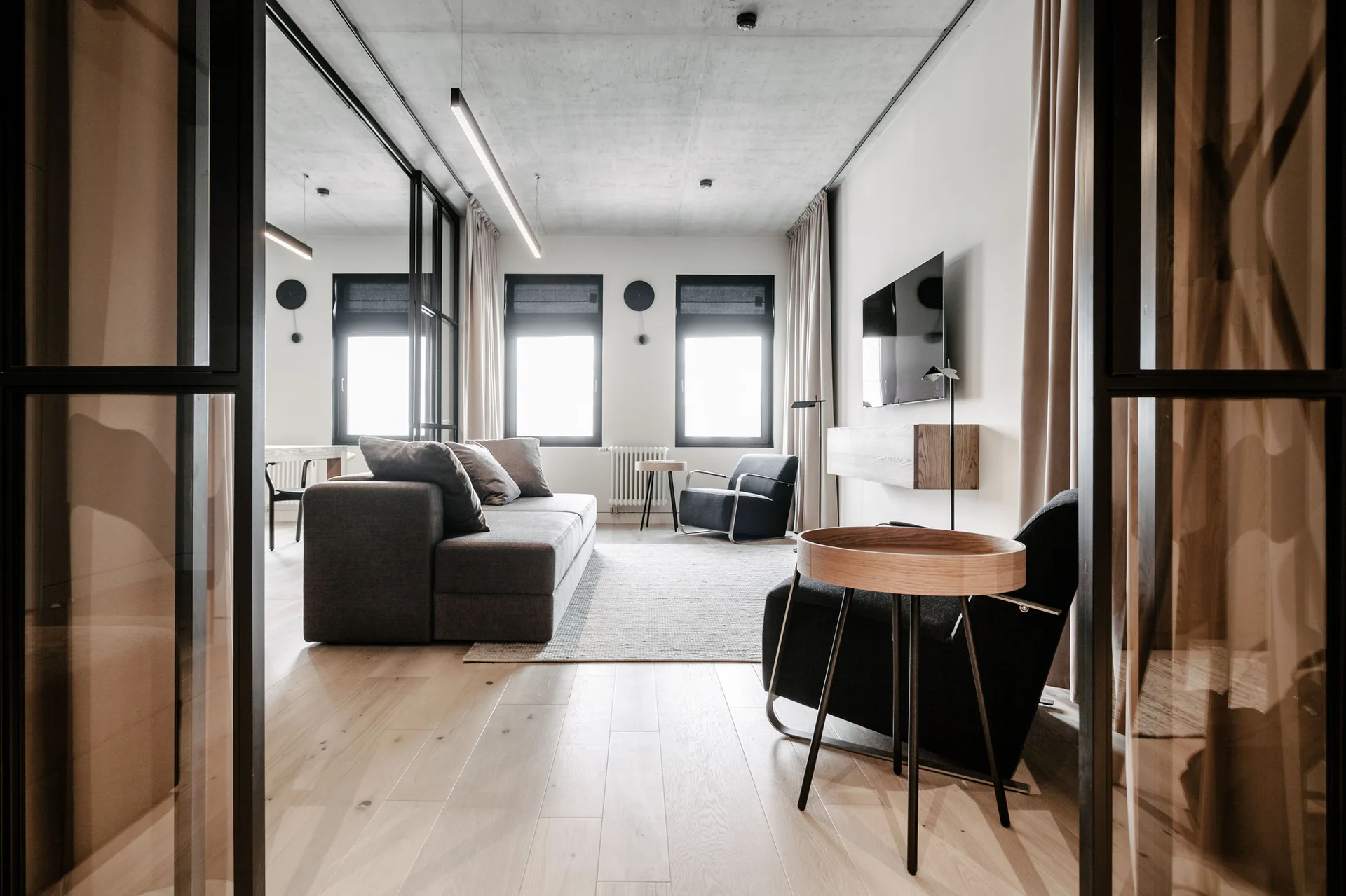 A modern living room features an exposed concrete ceiling, a grey sofa, a black-framed glass partition, and light wood flooring.