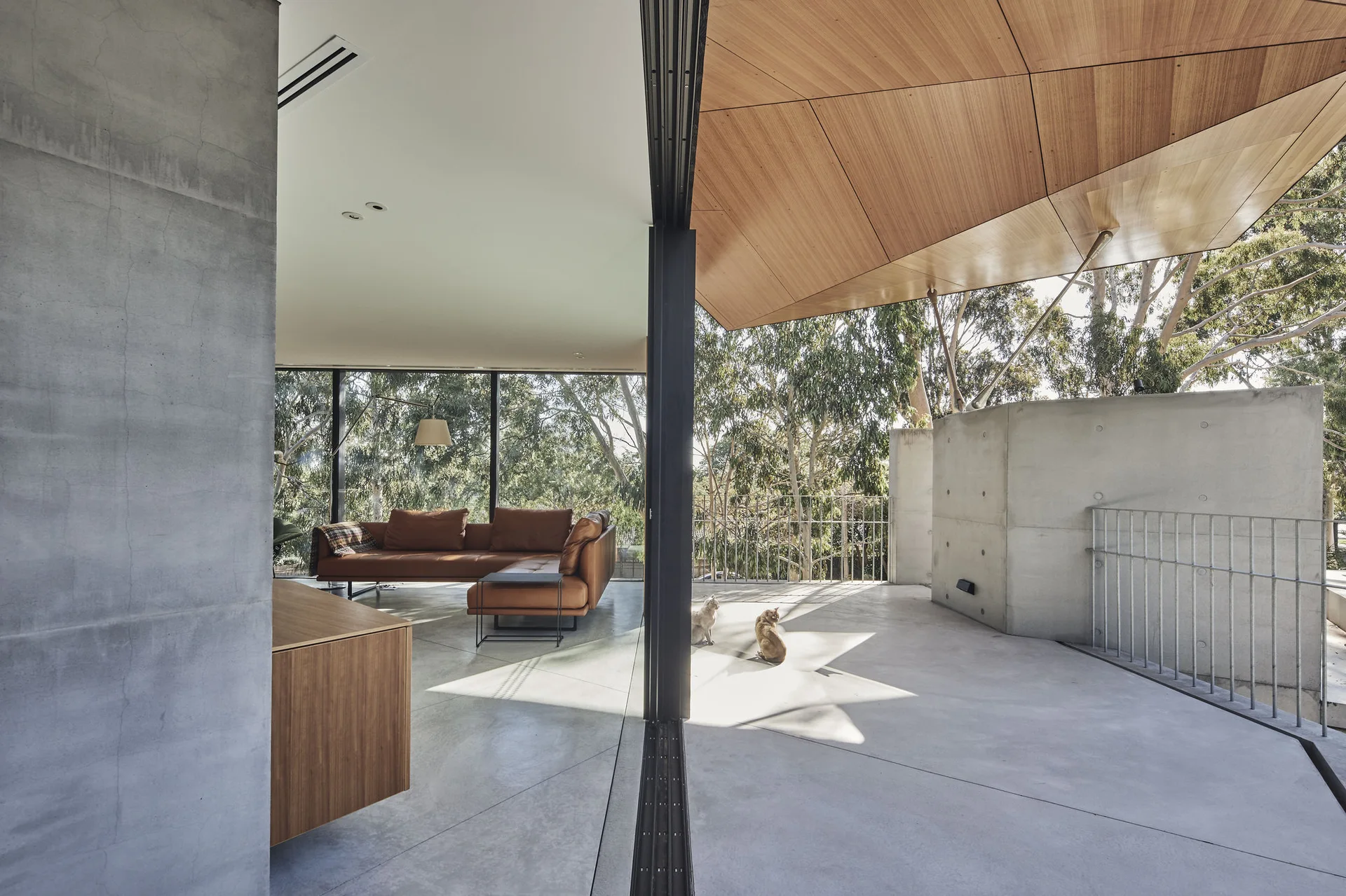 A modern living area with a brown leather sectional sofa opens onto a concrete terrace under an angled wood ceiling.