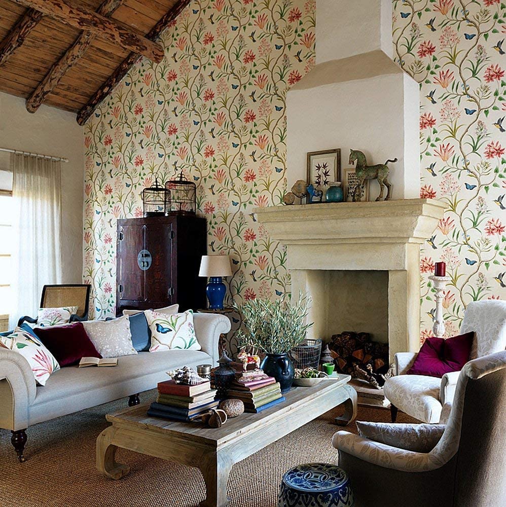 A rustic living room features botanical patterned wallpaper, exposed wooden ceiling beams, a stone fireplace, and a low wooden coffee table with books.