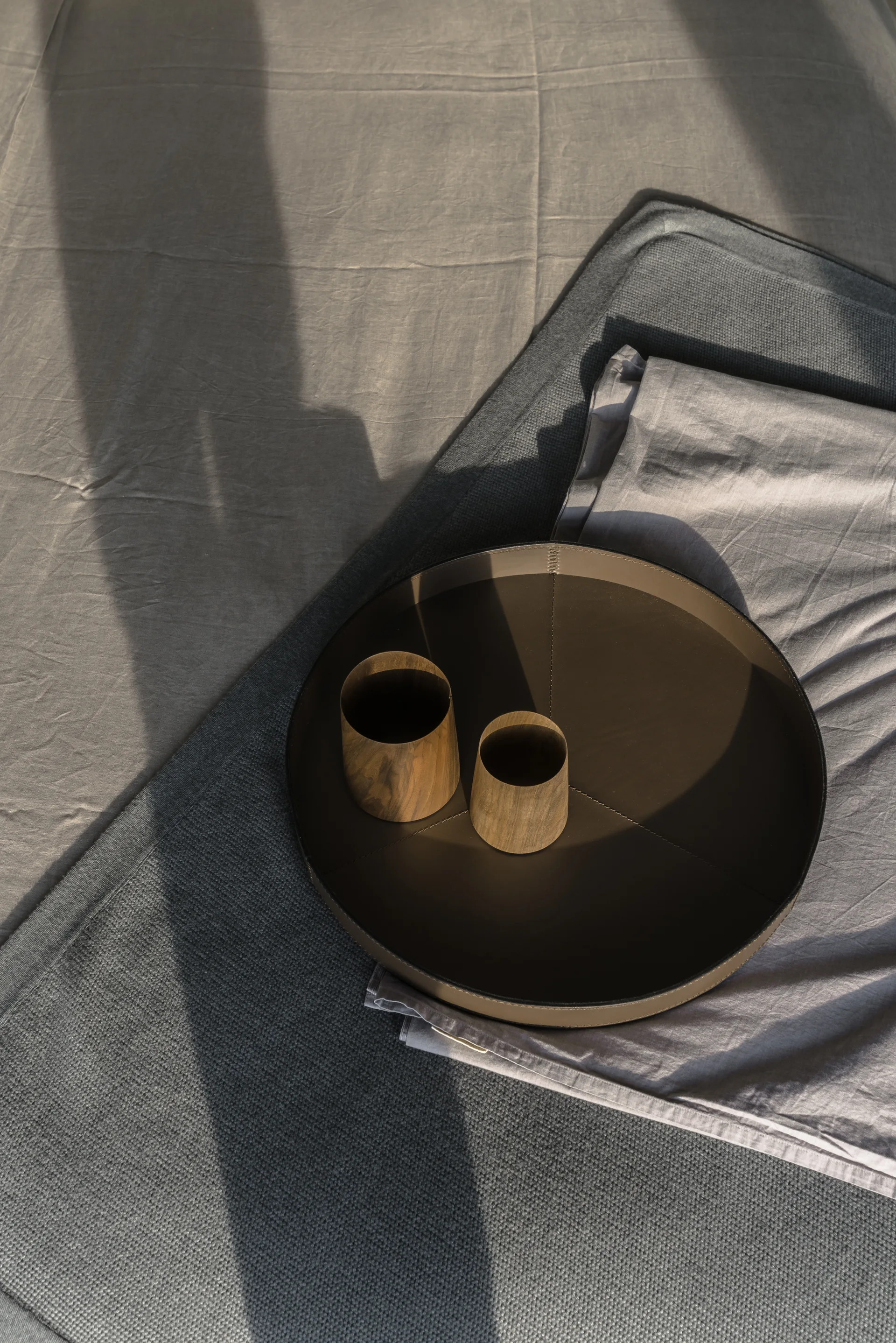 A dark brown circular leather tray holding two small wooden cups rests on layered gray bedding under strong natural shadows.
