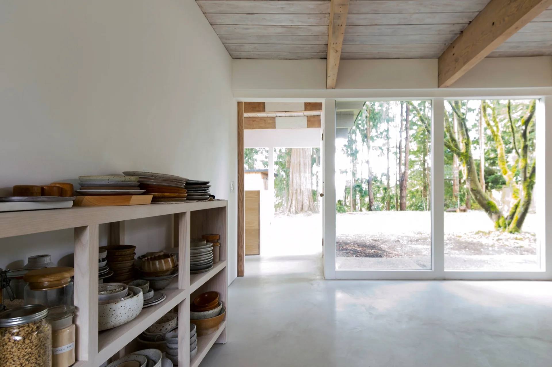 A low wooden shelving unit filled with ceramic tableware stands in a minimalist room with large windows showing a forest.