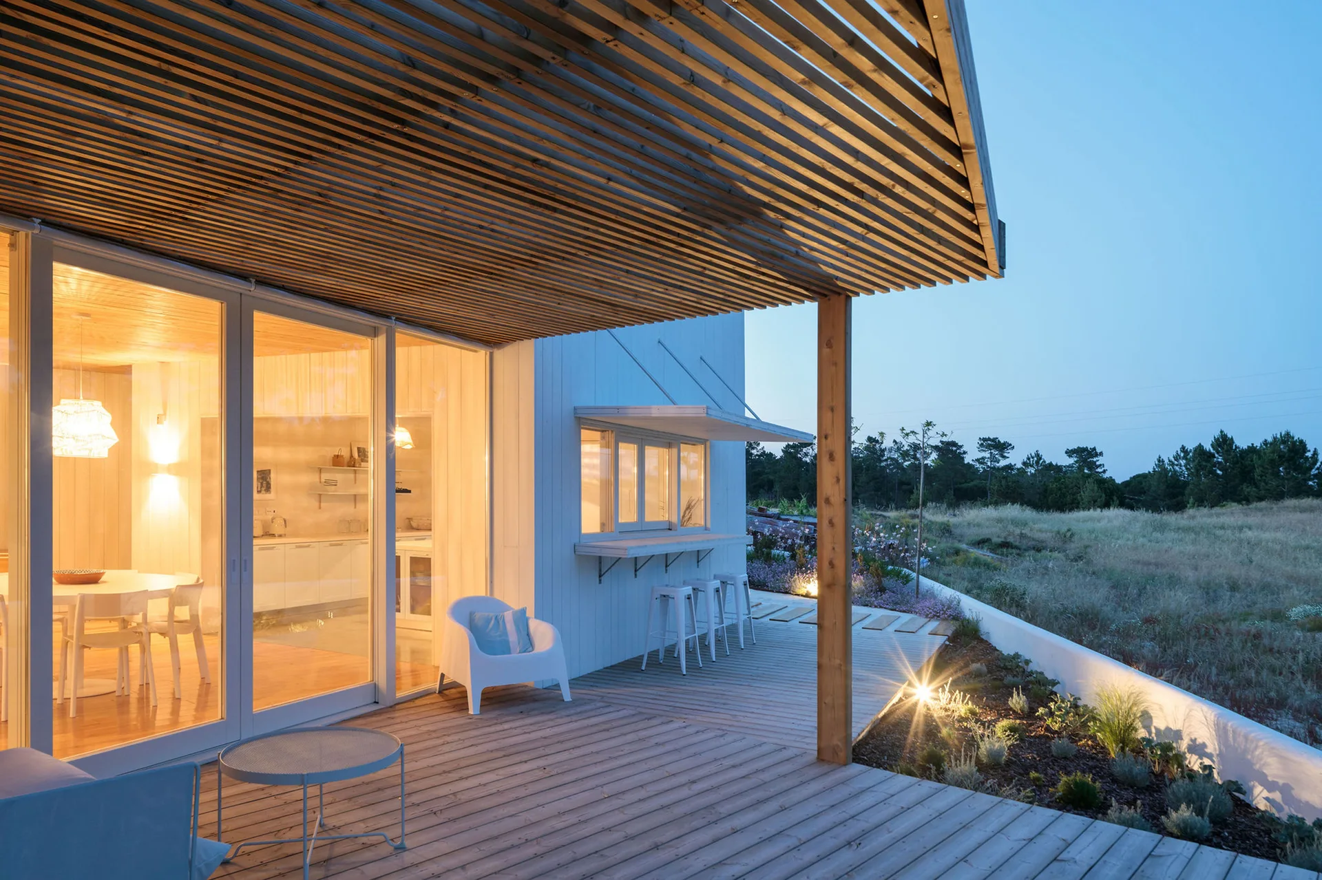 A wooden patio deck features a slatted timber pergola, a kitchen pass-through window with bar stools, and glowing glass sliding doors.