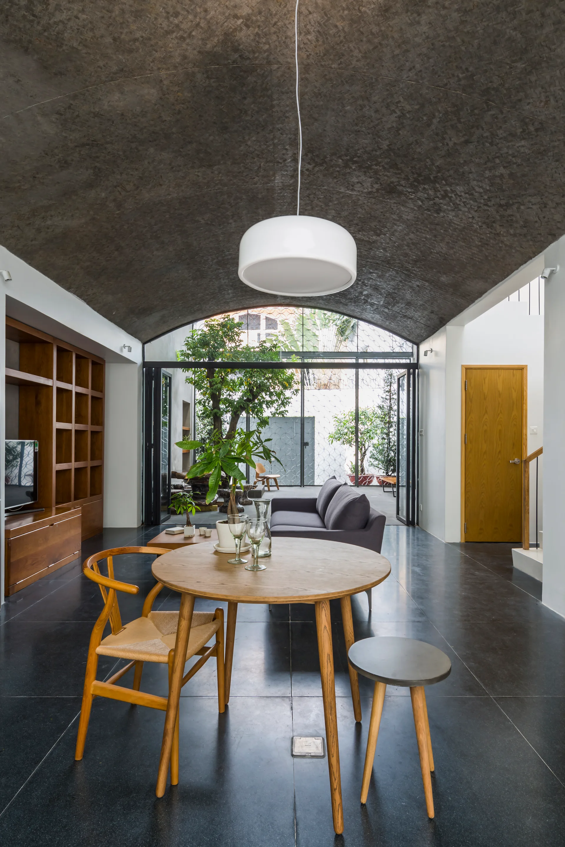 A modern dining room features a round wooden table, a dark textured vaulted ceiling, and glass folding doors leading to an outdoor courtyard.