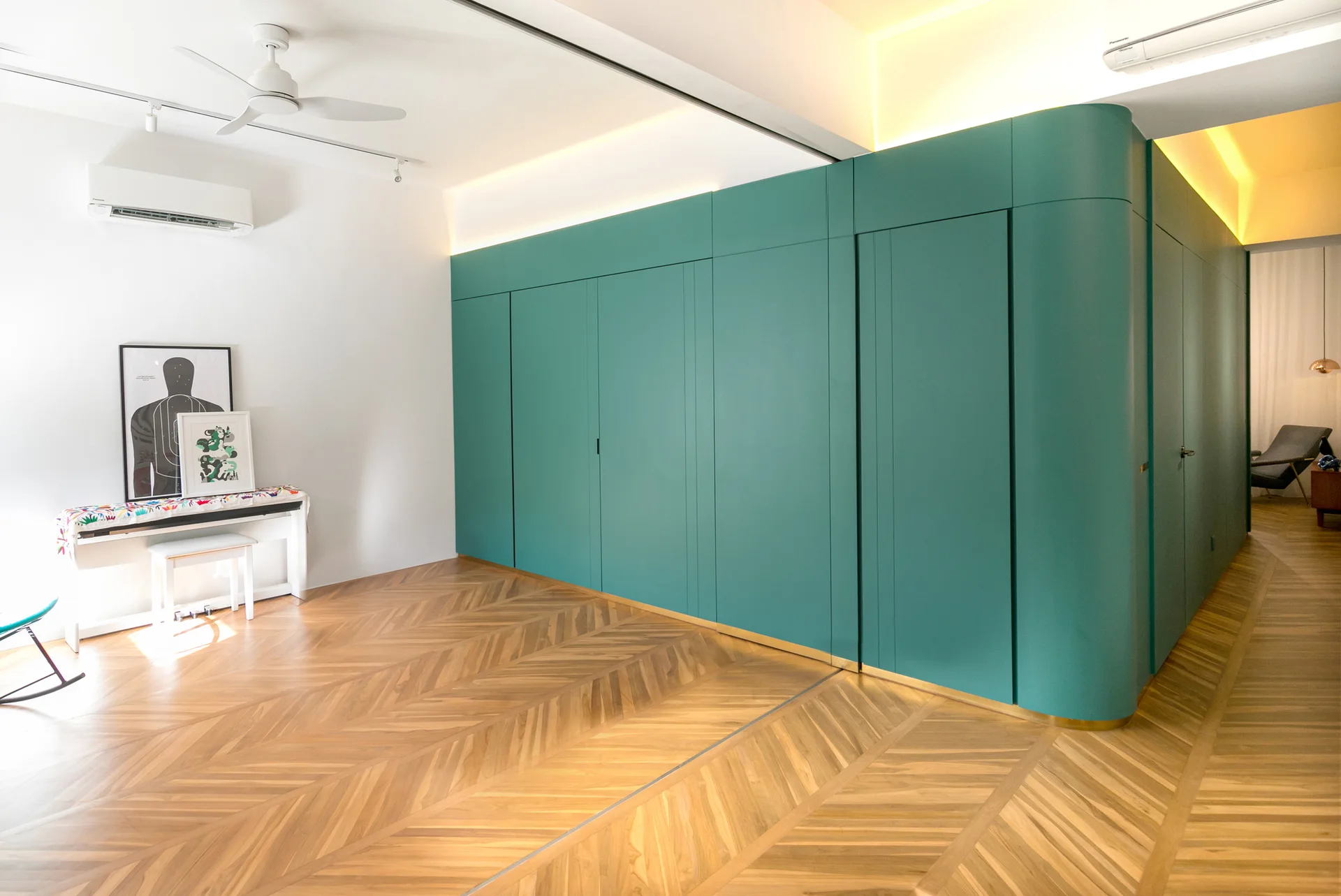 A modern living area with a large curved teal storage wall, cove lighting, and chevron pattern wood flooring.