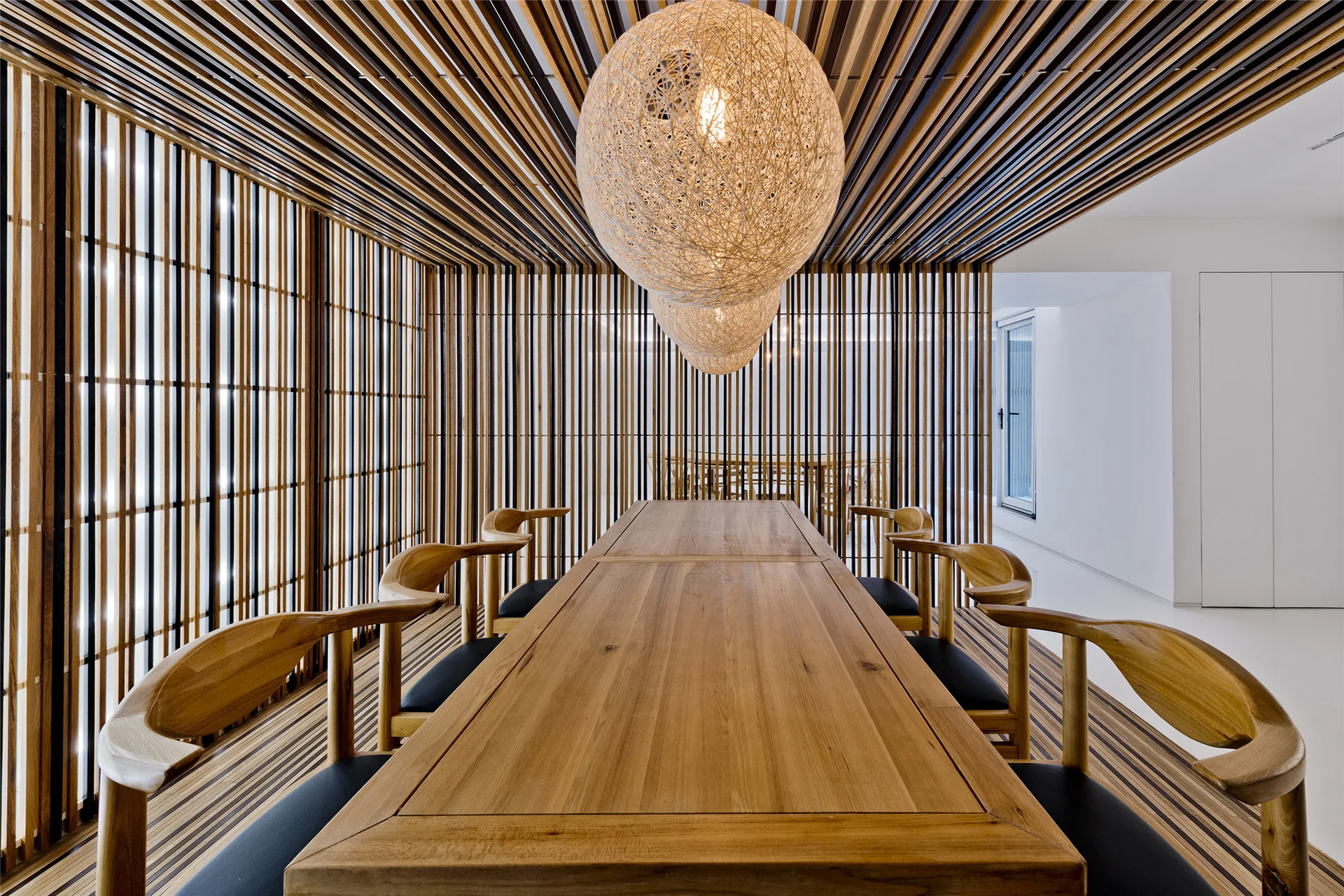 A long wooden dining table with curved wood chairs sits inside a room wrapped in vertical wood slats under spherical woven pendant lights.