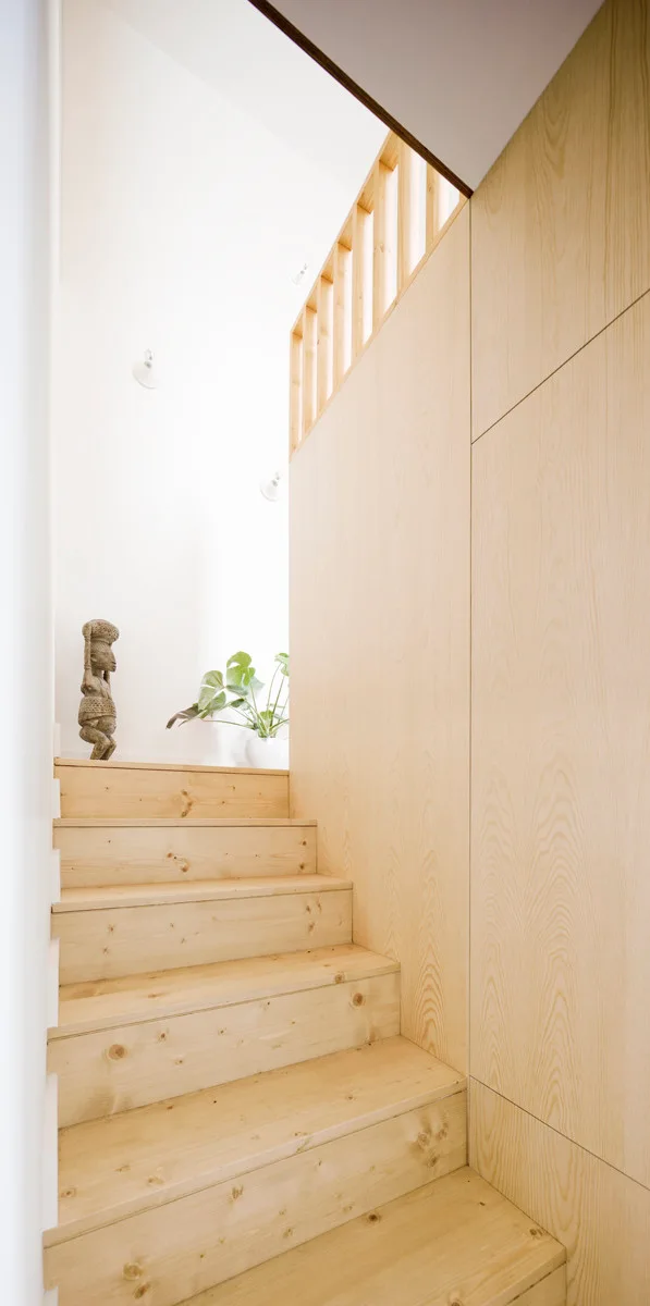 A light wood staircase with natural wood grain steps leads up past a large plywood-paneled wall and a simple slatted wooden railing.