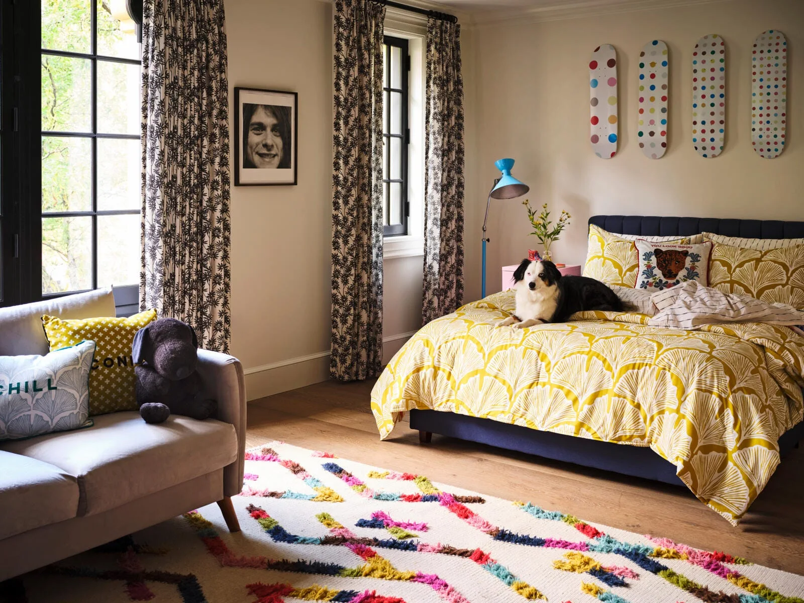 A playful bedroom features a blue upholstered bed with yellow bedding, patterned curtains, a colorful rug, and skateboards mounted on the wall.