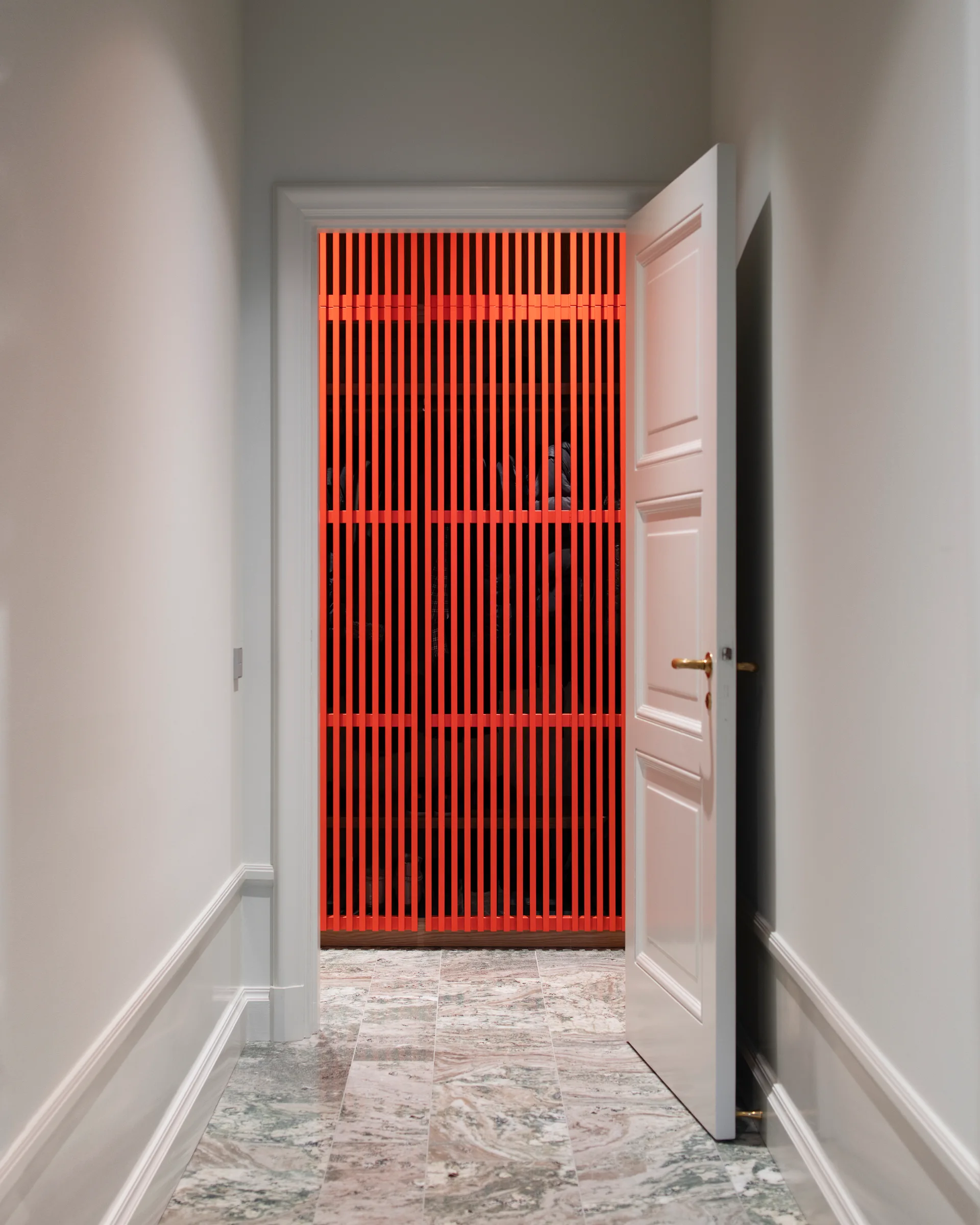 An open white paneled door reveals a closet lined with bright orange vertical wooden slats, set against a grey-and-white marble hallway floor.