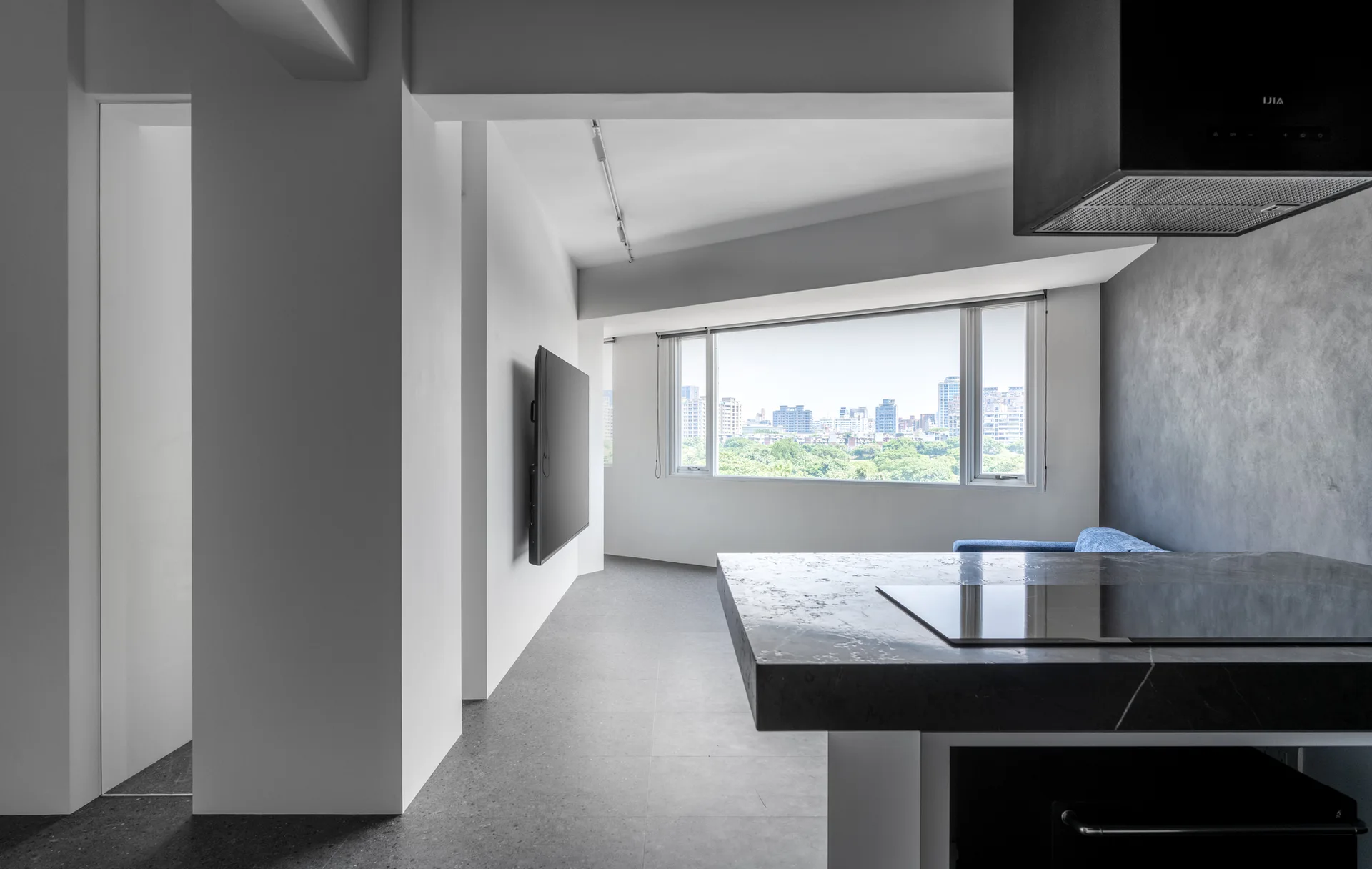 A minimalist open kitchen features a dark stone island with a cooktop, a wall-mounted television, and a large window overlooking a cityscape.