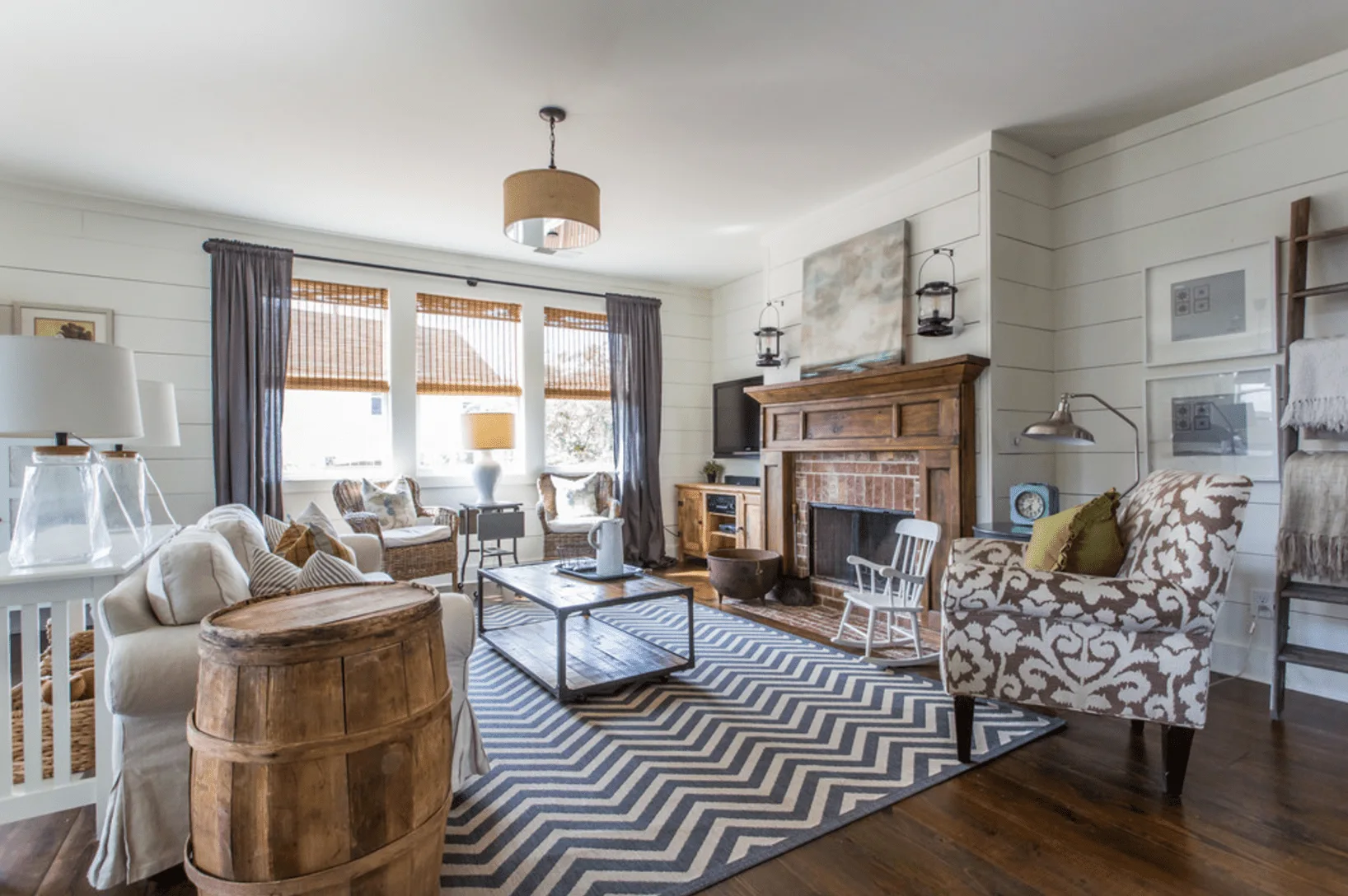 A farmhouse living room features white shiplap walls, a central brick fireplace with a wood mantle, a patterned armchair, and a chevron rug.