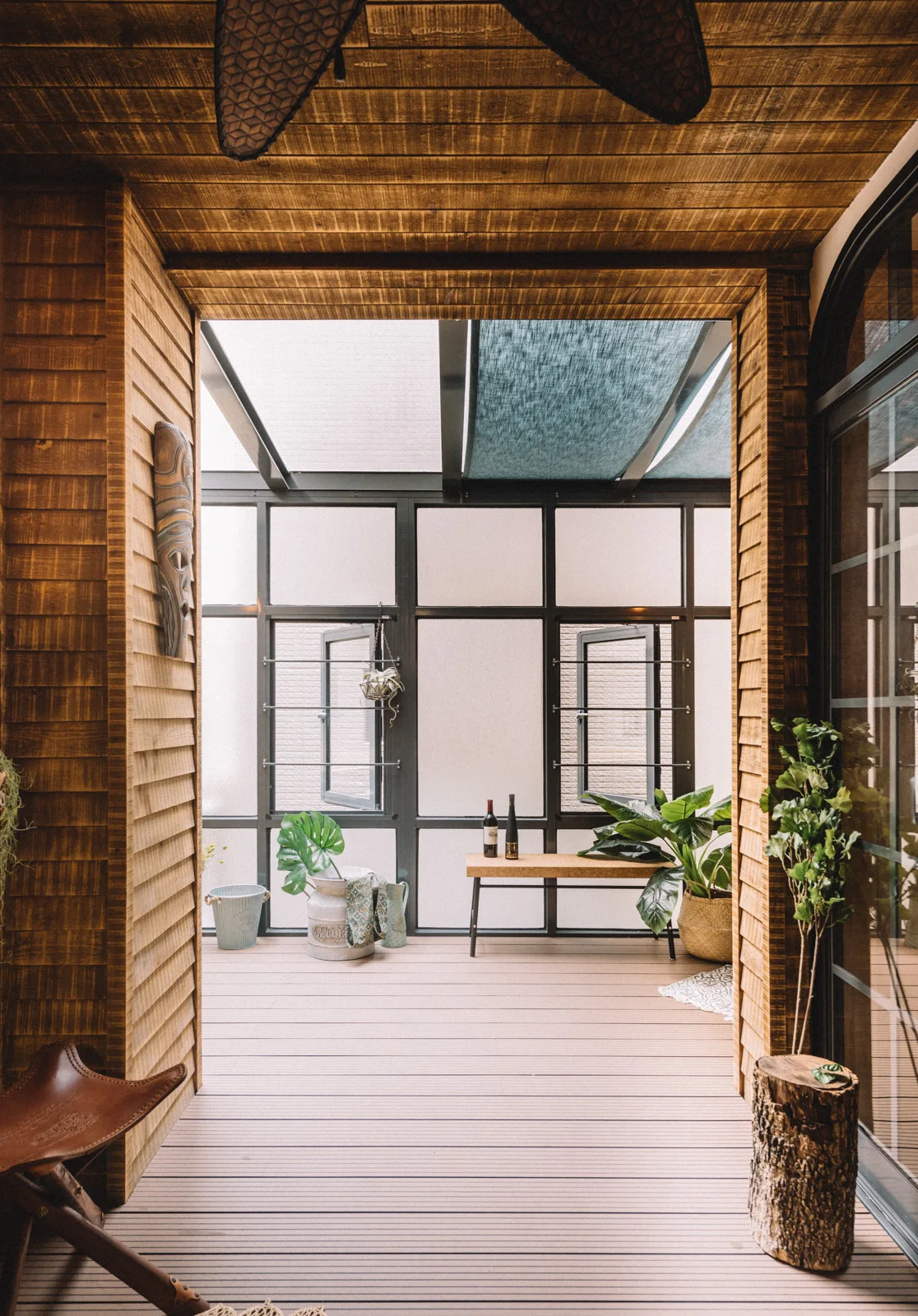 A wood-paneled sunroom features a light-colored wood floor, a simple wooden bench, potted plants, and a wall of black-framed windows.