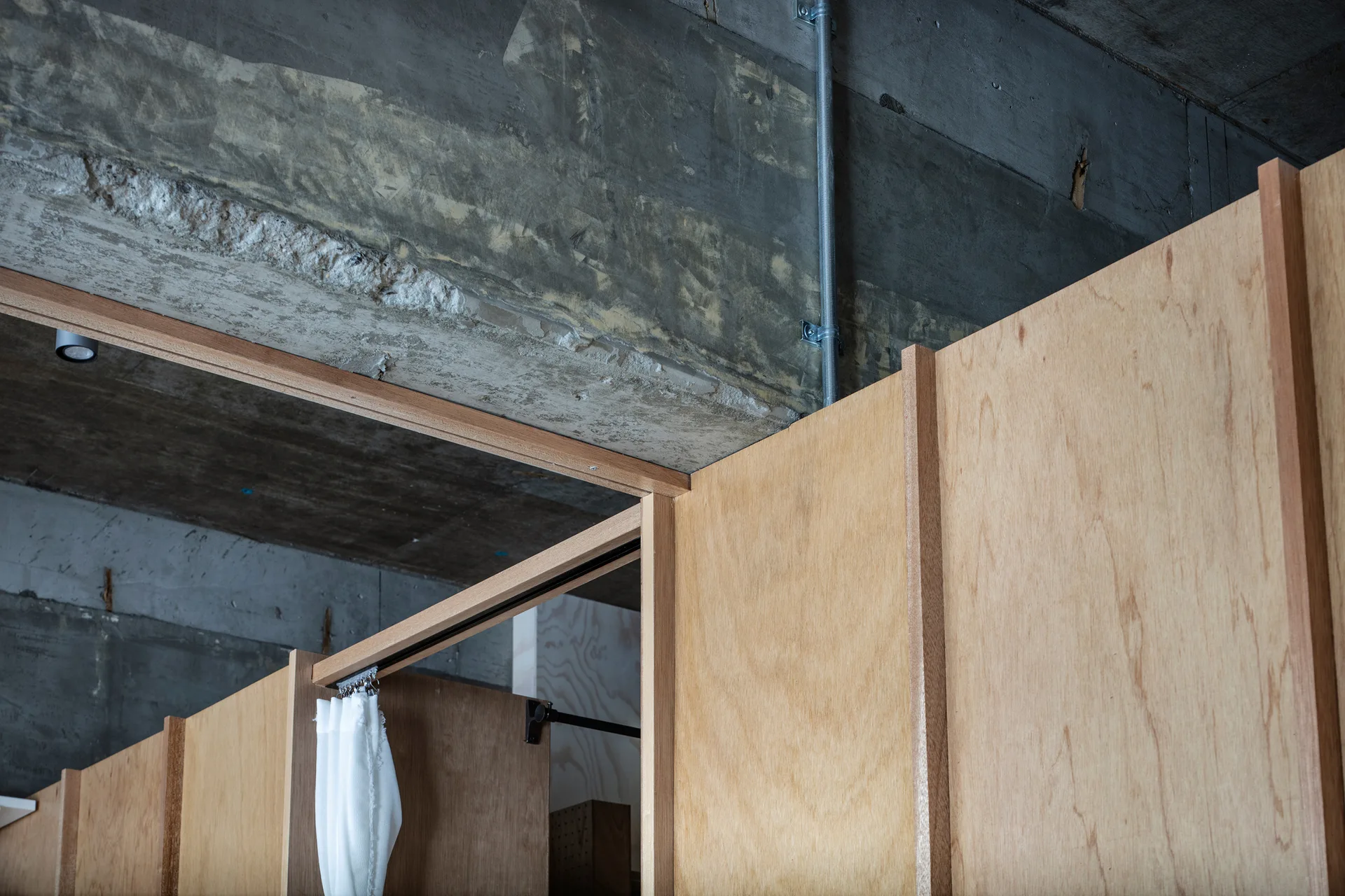 A close-up view of a light plywood partition wall with vertical wood trim meeting a rough, gray concrete ceiling with a metal conduit pipe.