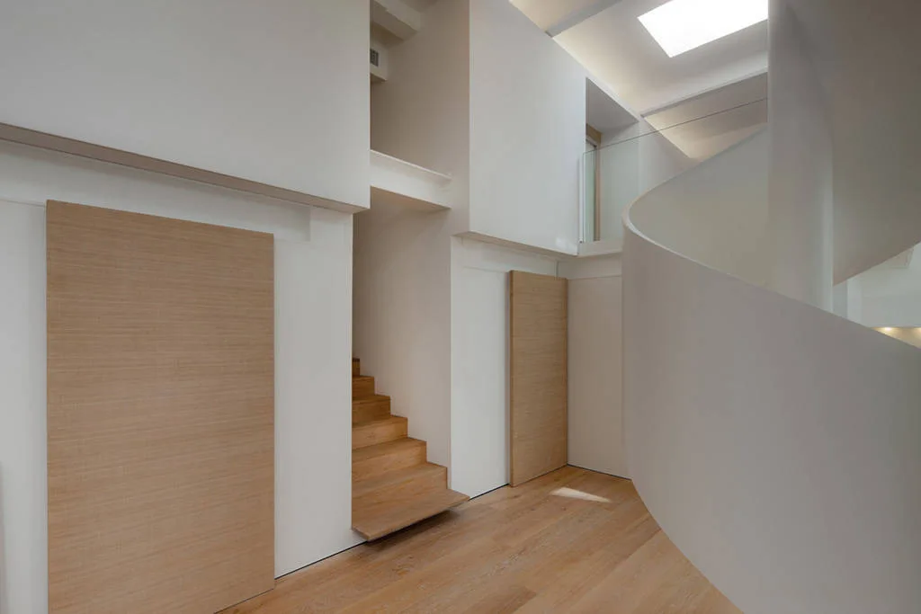 A minimalist white foyer featuring a sculptural spiral staircase, light wood floors, and two rectangular wood wall panels.