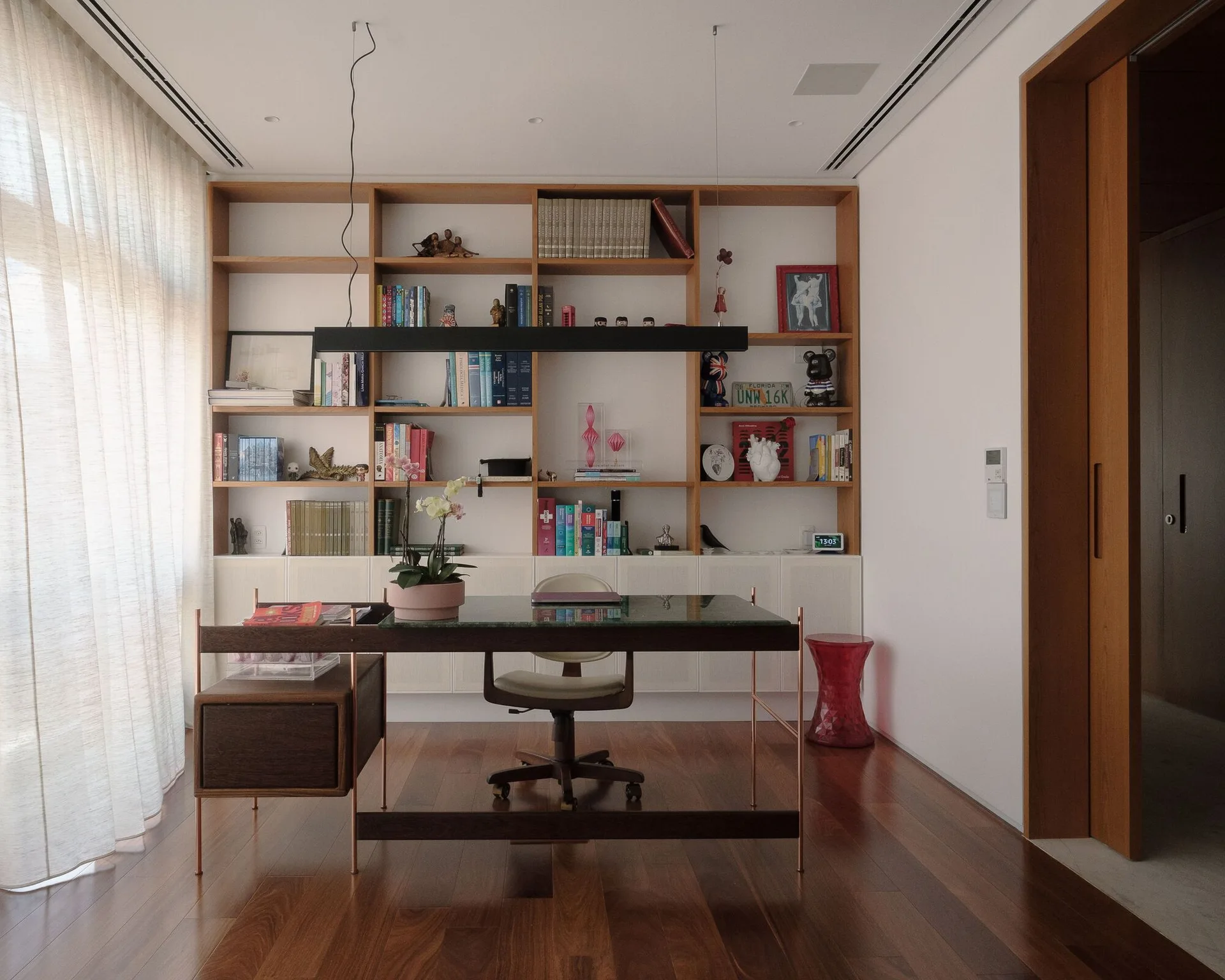 A modern home office featuring a dark wood desk with a glass top, a large built-in wooden shelving unit, and sheer white curtains.