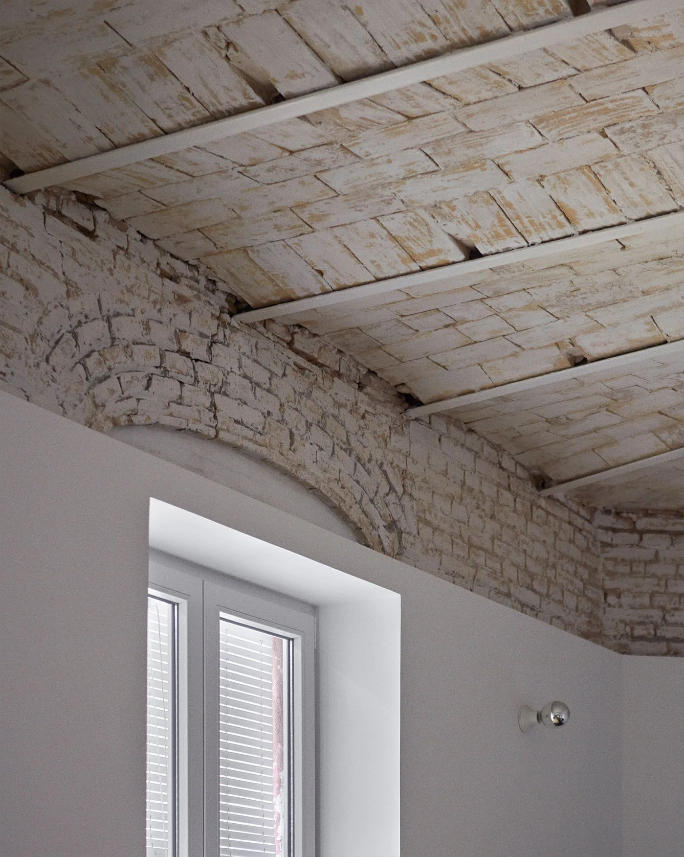 A vaulted brick ceiling meets a smooth white plaster wall featuring a recessed window with blinds and a metallic spherical sconce.