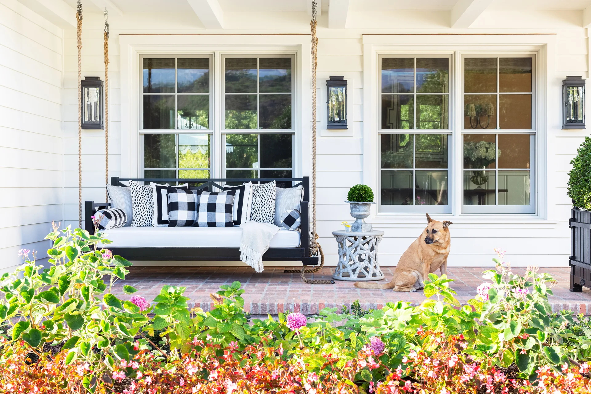 A black hanging porch swing with black and white patterned cushions rests on a red brick floor next to a carved grey side table.
