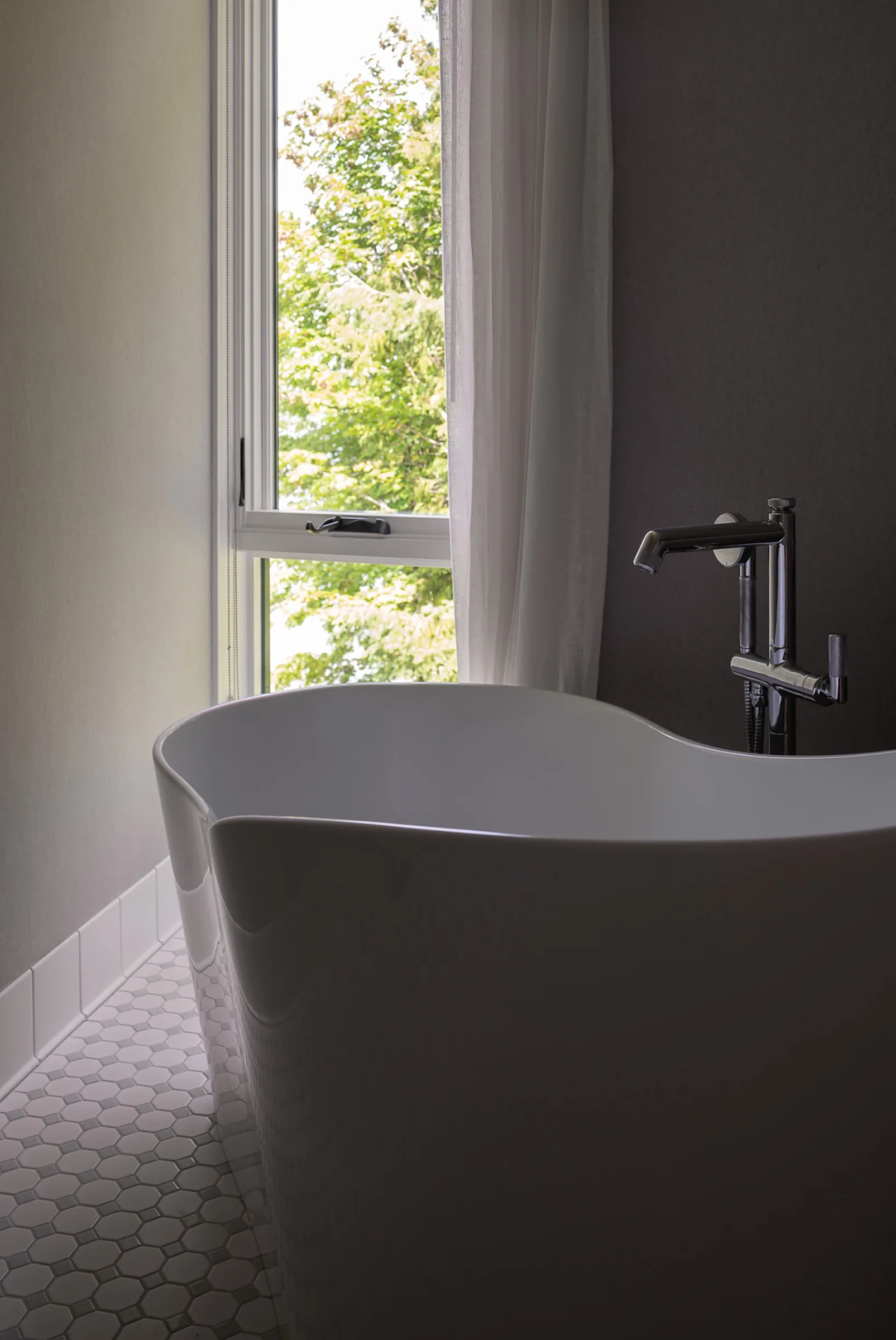 A white freestanding soaking tub sits on a grey hexagonal tile floor next to a window with a sheer white curtain.