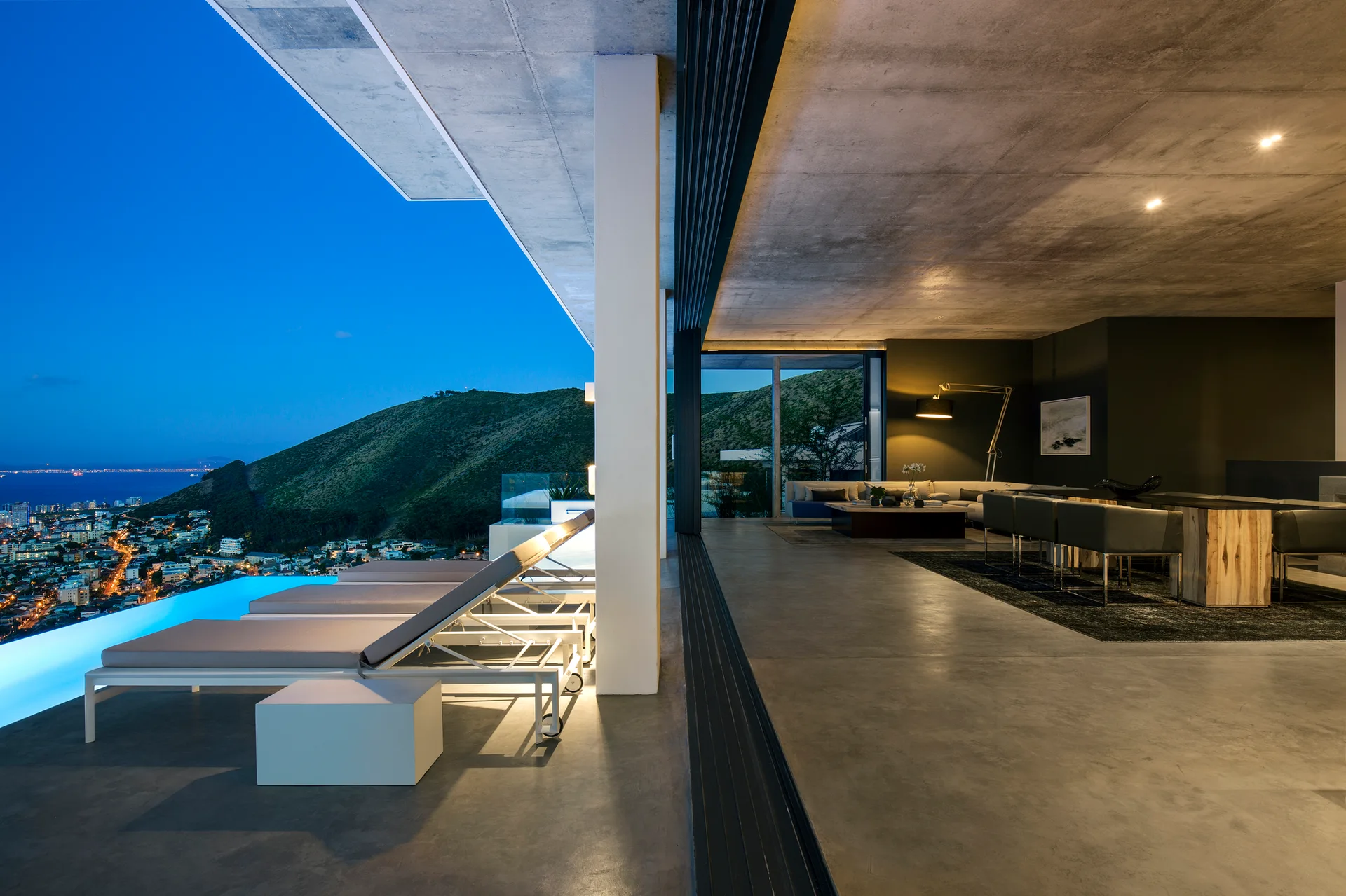 A modern patio with concrete floors and lounge chairs next to an illuminated infinity pool overlooking a city landscape at dusk.