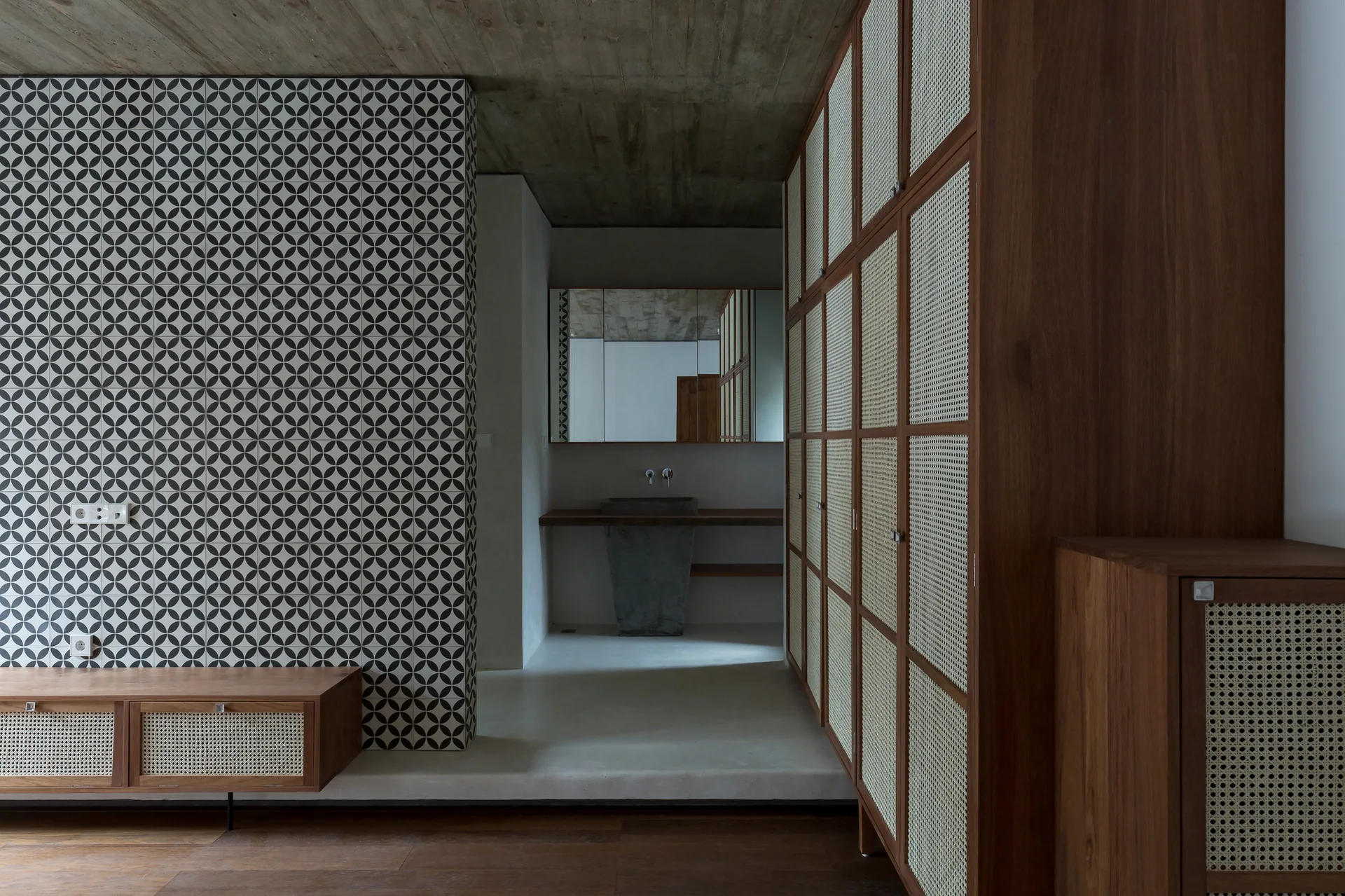 A modern bathroom entryway features tall wooden cabinets with woven cane panels, an exposed concrete ceiling, and a black-and-white patterned tile wall.