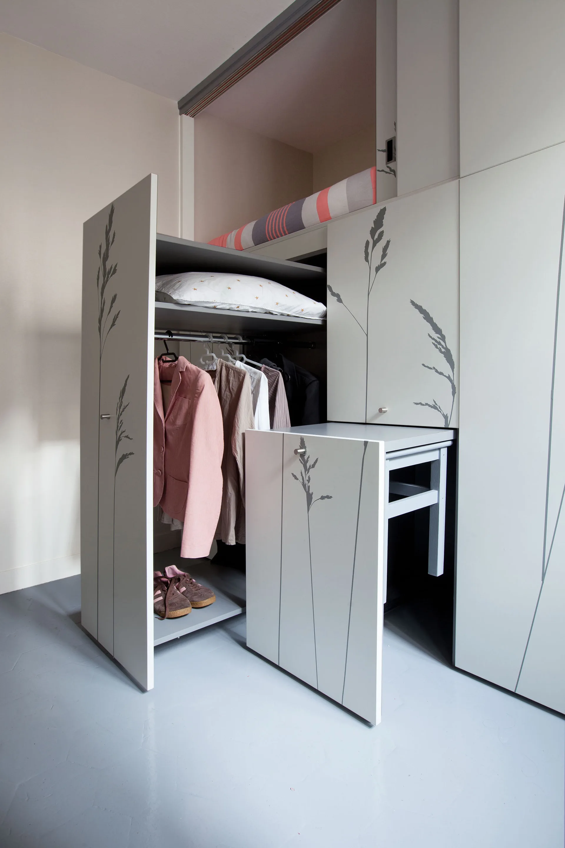 A custom off-white closet unit features doors with botanical line art graphics, showing a hanging clothes rack and an integrated pull-out desk.