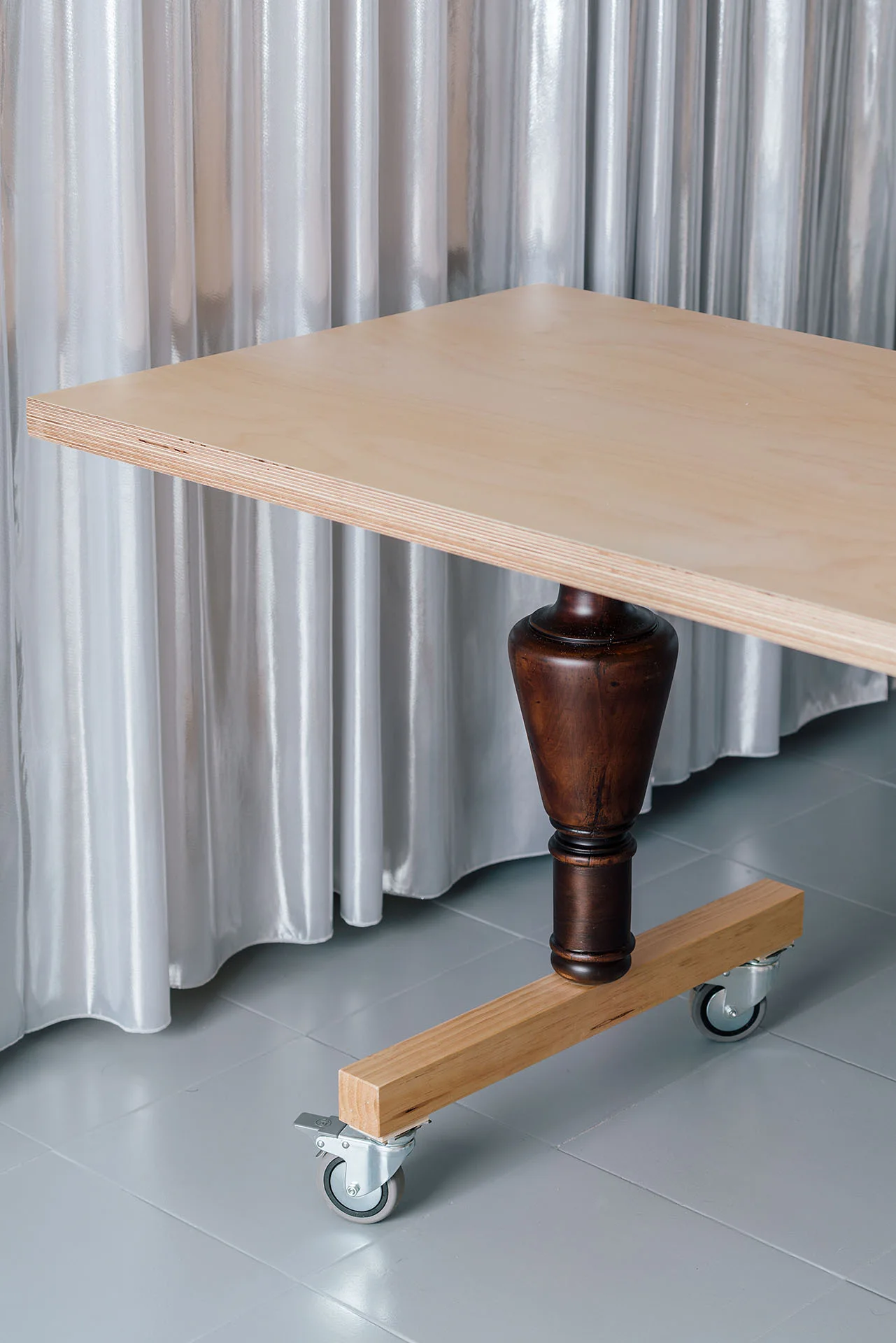A light wood table with a dark turned wooden pedestal leg and metal casters stands on a grey tile floor against a silver curtain background.