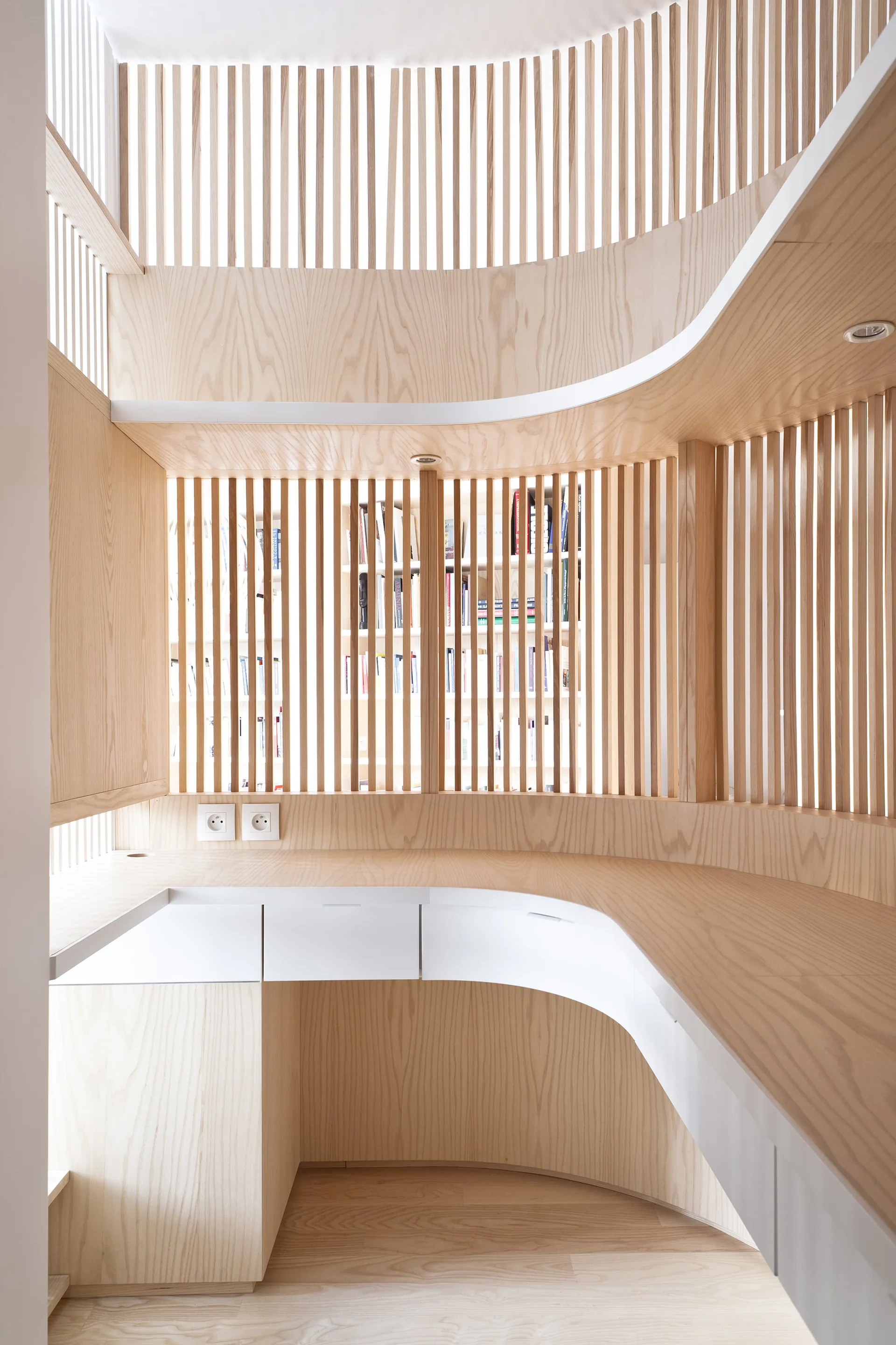 A curved light wood built-in desk with white drawer fronts is positioned under a vertical wooden slat screen that reveals a bookshelf behind it.