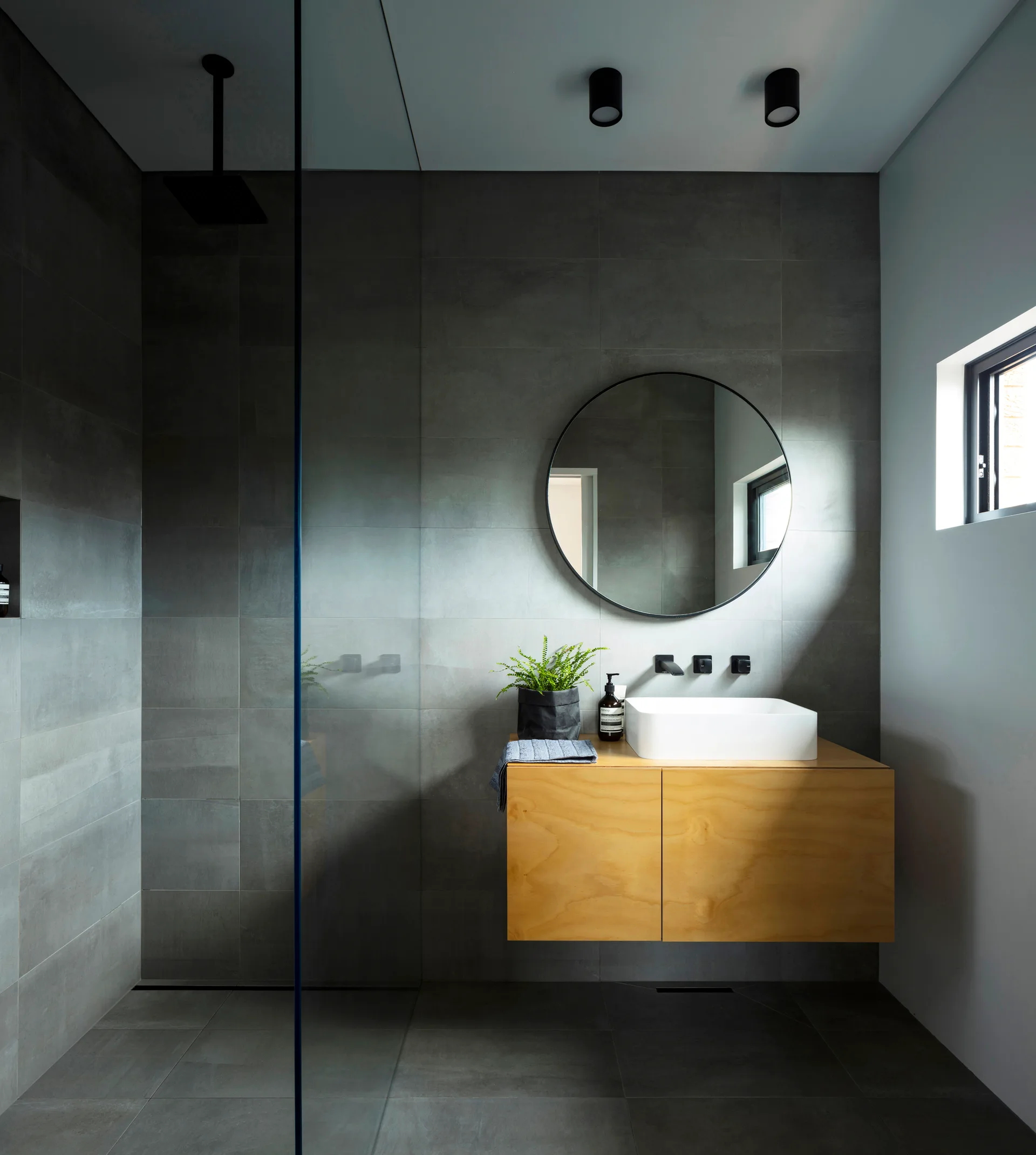 A minimalist bathroom featuring large-format grey wall tiles, a floating wood-grain vanity with a white vessel sink, and a round black-framed mirror.