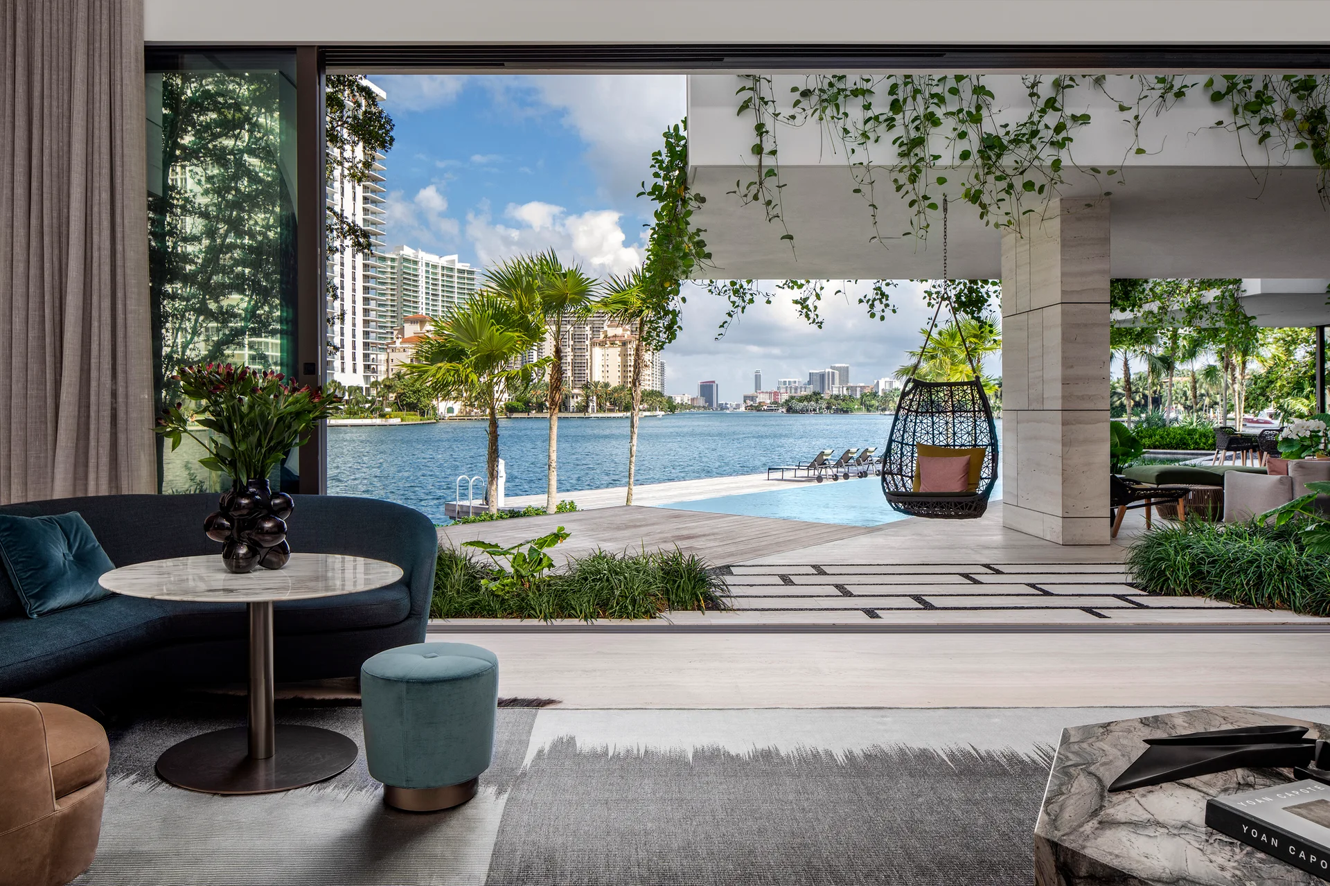 A contemporary living room features a curved blue sofa and marble pedestal table next to a retractable glass wall opening onto a waterfront patio.