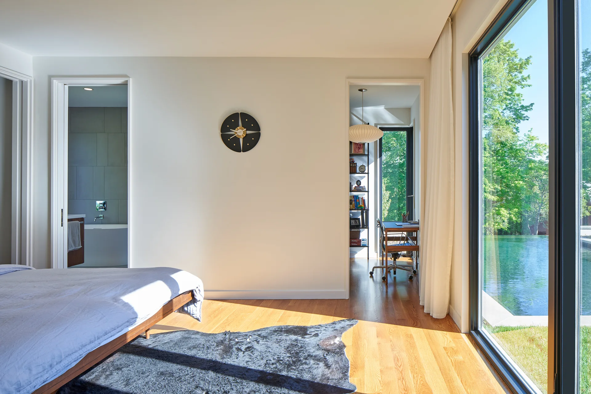 A sunlit modern bedroom with wood flooring, an animal hide rug, and floor-to-ceiling glass windows overlooking a pool.