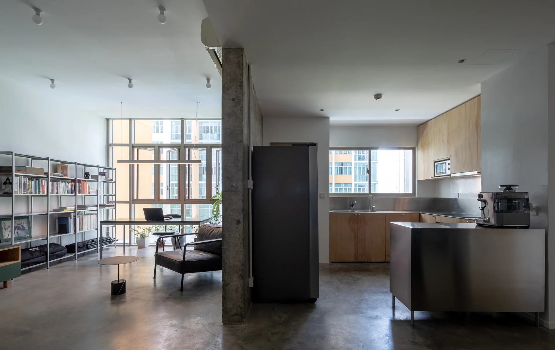 An industrial-style open-plan apartment features a stainless steel kitchen island, light wood cabinetry, and an exposed concrete pillar beside a living area.