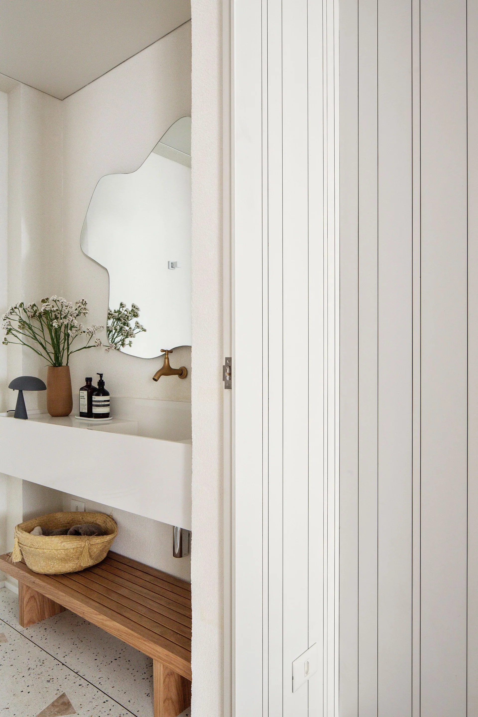 A minimalist bathroom vanity area features an organic-shaped mirror, a white floating sink with a brass faucet, and a slatted wood bench.