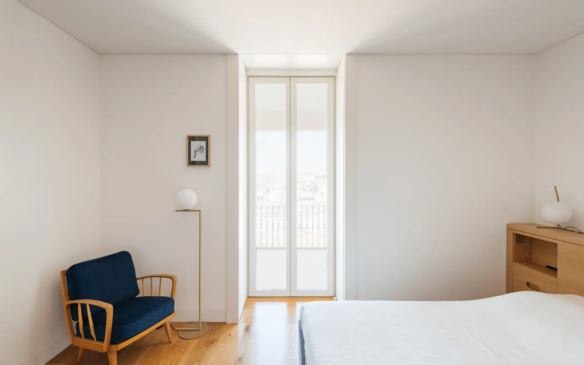 A minimalist white bedroom featuring a deep blue upholstered armchair, a tall floor lamp with an orb shade, and a light wood headboard with integrated shelves.