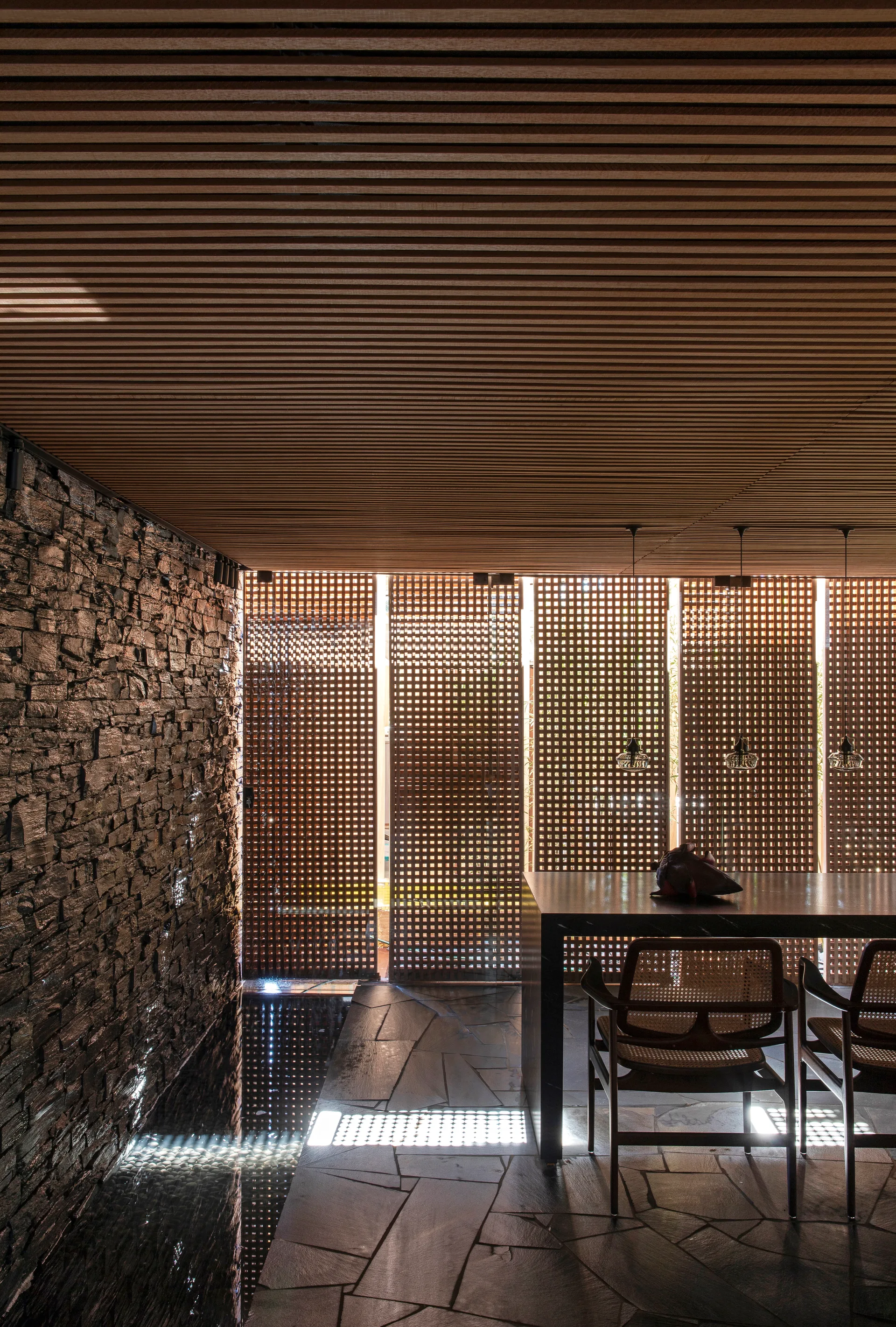 A modern dining room featuring a slatted wood ceiling, a rugged stone wall with a water feature, and grid-patterned wood privacy screens.