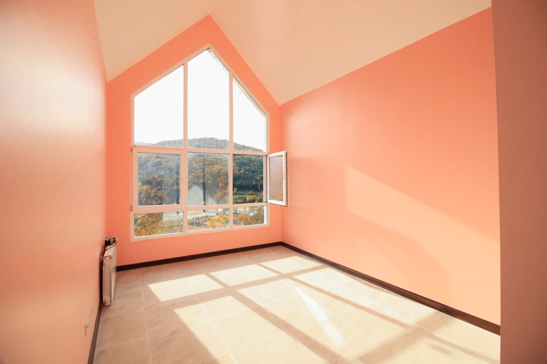 An empty attic room features peach painted walls, a vaulted ceiling, a tiled floor, and a large gabled window showing a mountain view.