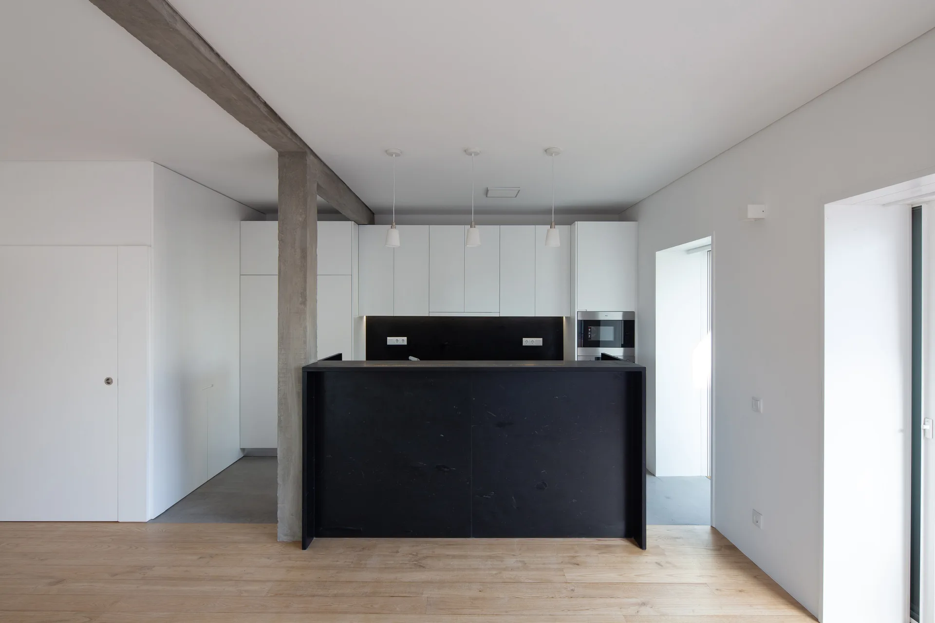 A minimalist kitchen features a matte black island, white flat-front cabinets, three pendant lights, and an exposed raw concrete column on wood flooring.
