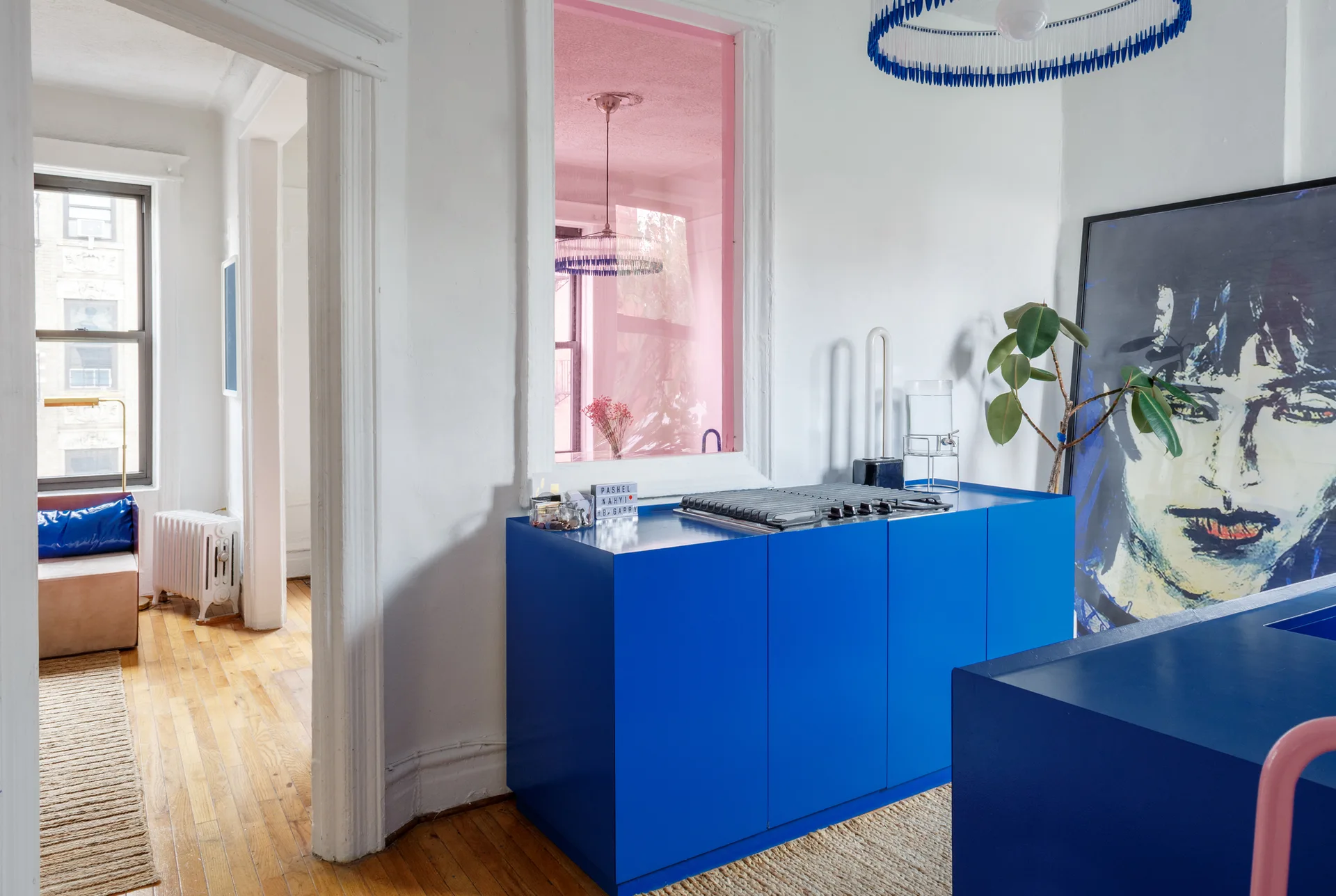 A modern kitchen features vibrant cobalt blue cabinets, a pink-tinted interior window, and natural hardwood flooring against white walls.