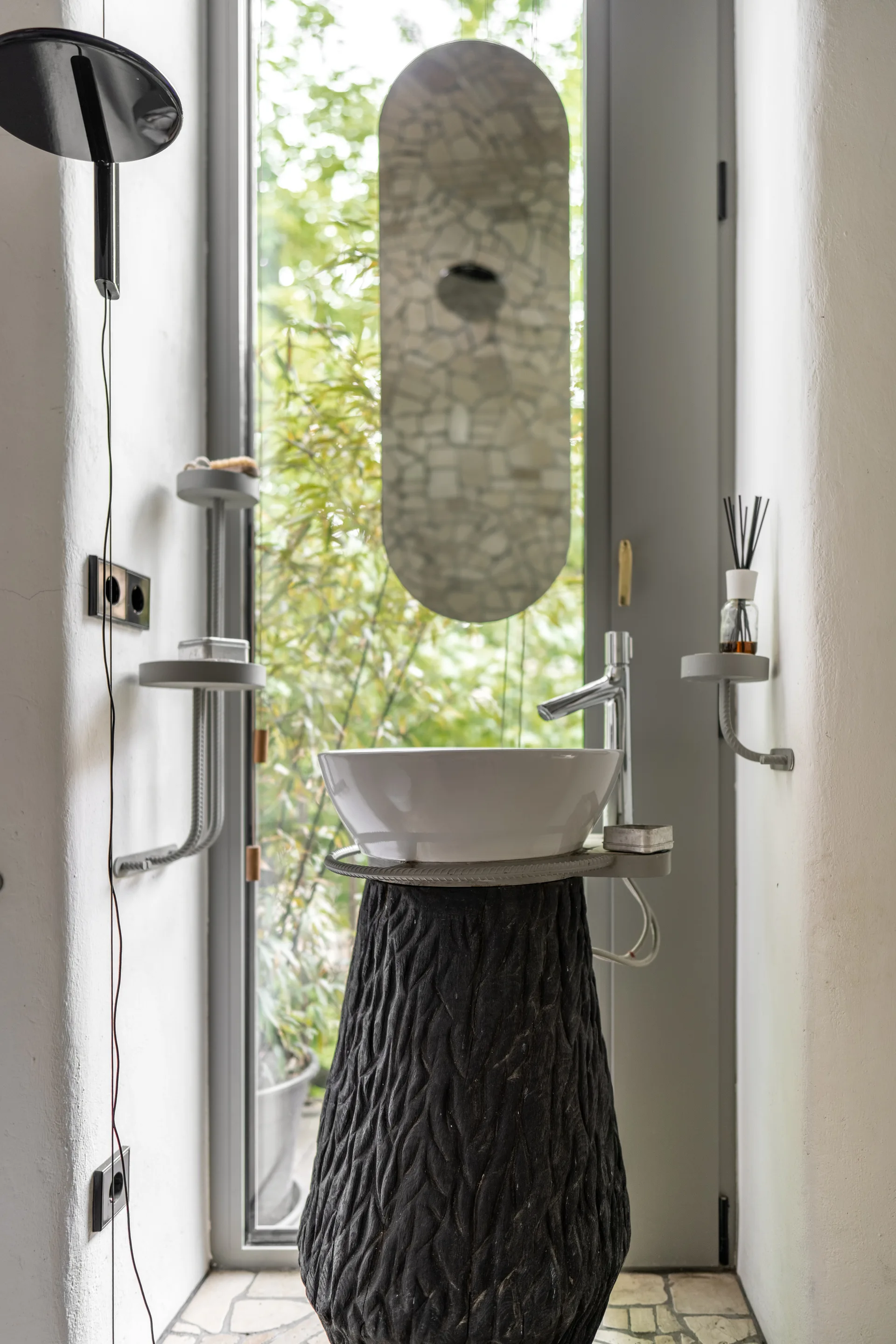 A sculptural pedestal sink with a white ceramic basin and textured black base sits in front of a tall glass door with an oval mirror.